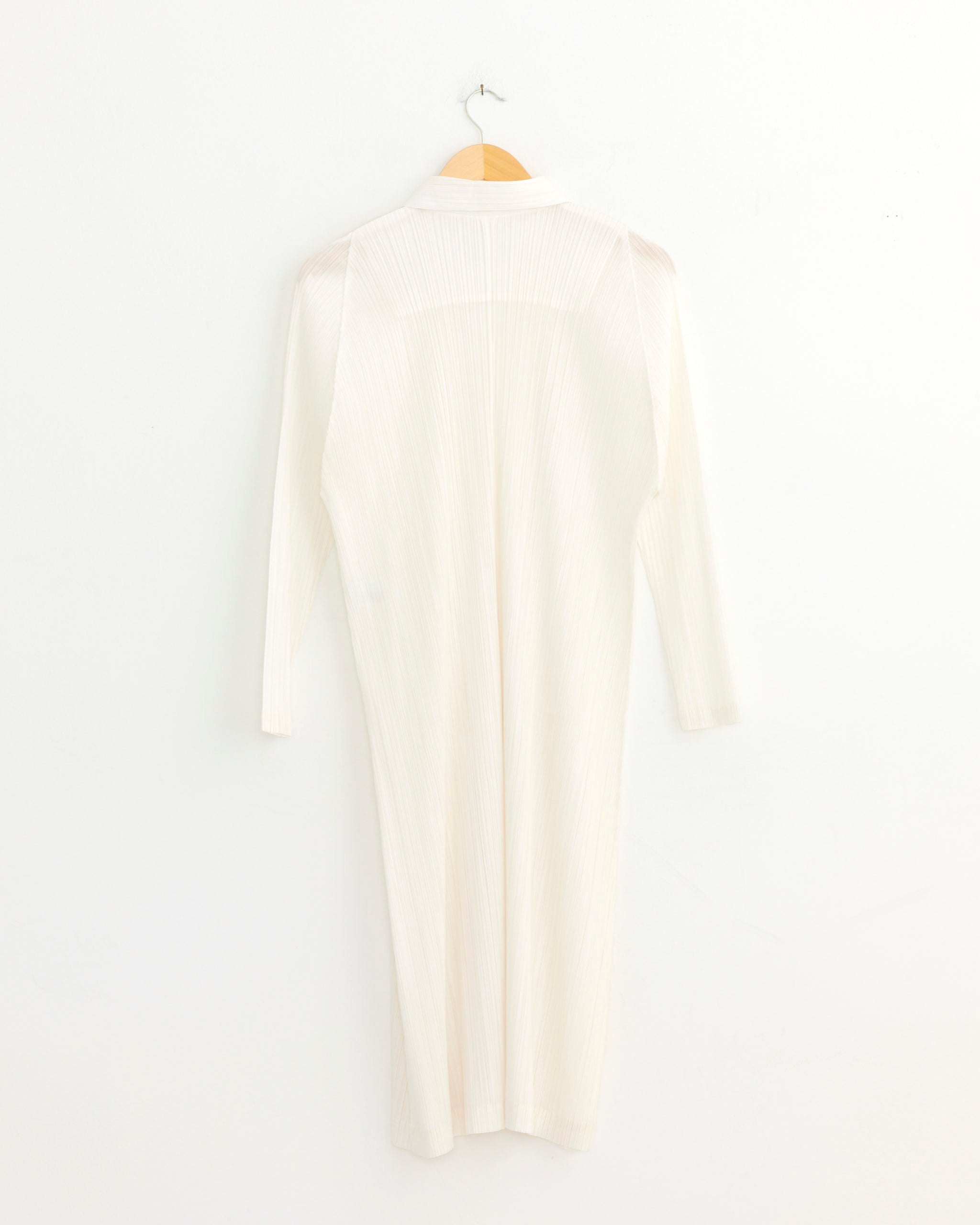 The MC Feb Coat in White by Pleats Please Issey Miyake, a long-sleeved, ankle-length dress with a collar and vertical ribbed texture, hangs on a wooden hanger against a plain white wall.