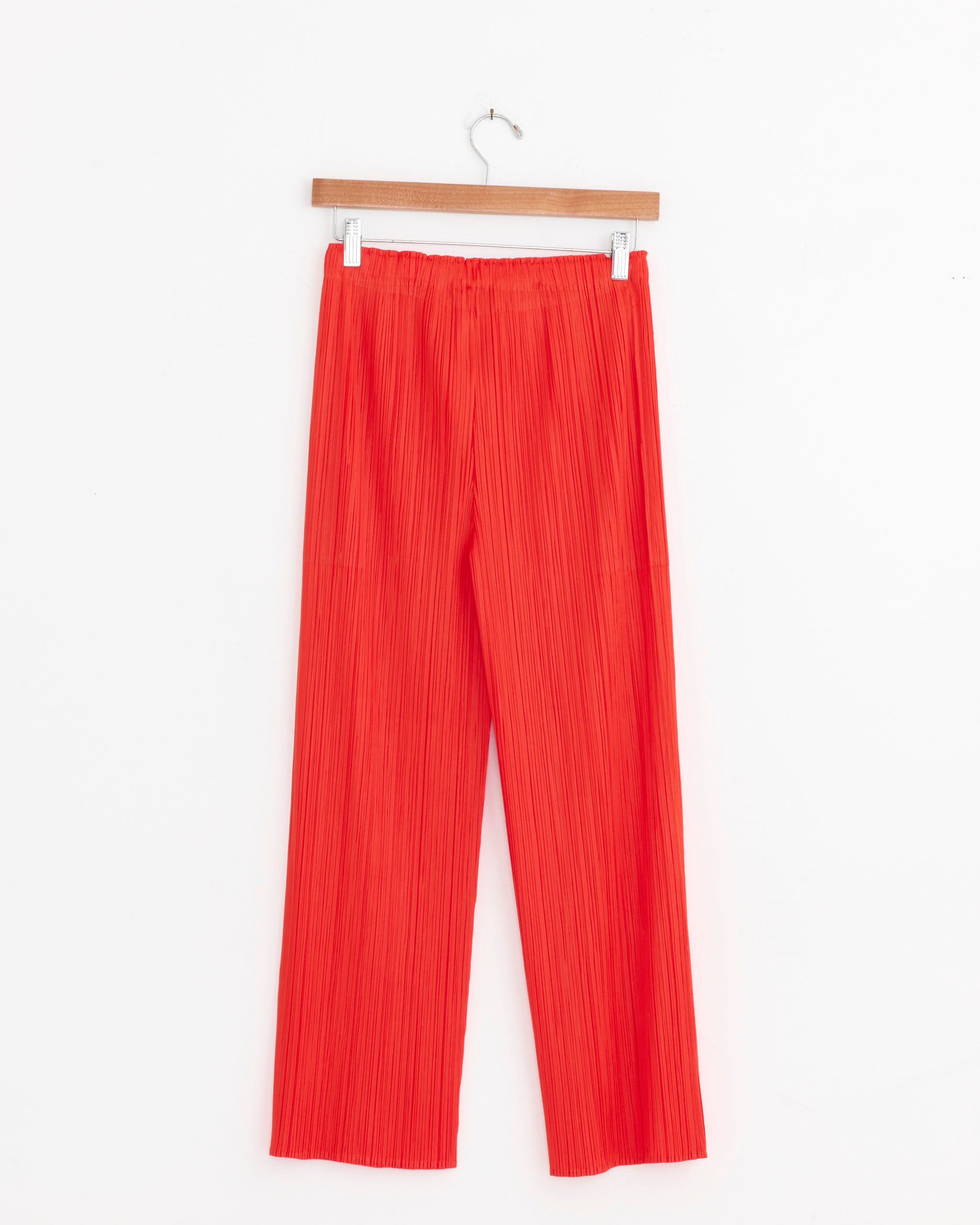 The MC Feb Pant in Red by Pleats Please Issey Miyake features bright red pleated straight-leg design, shown on a wooden hanger against a white background. For sizing or fit questions, please use our online chat assistance.
