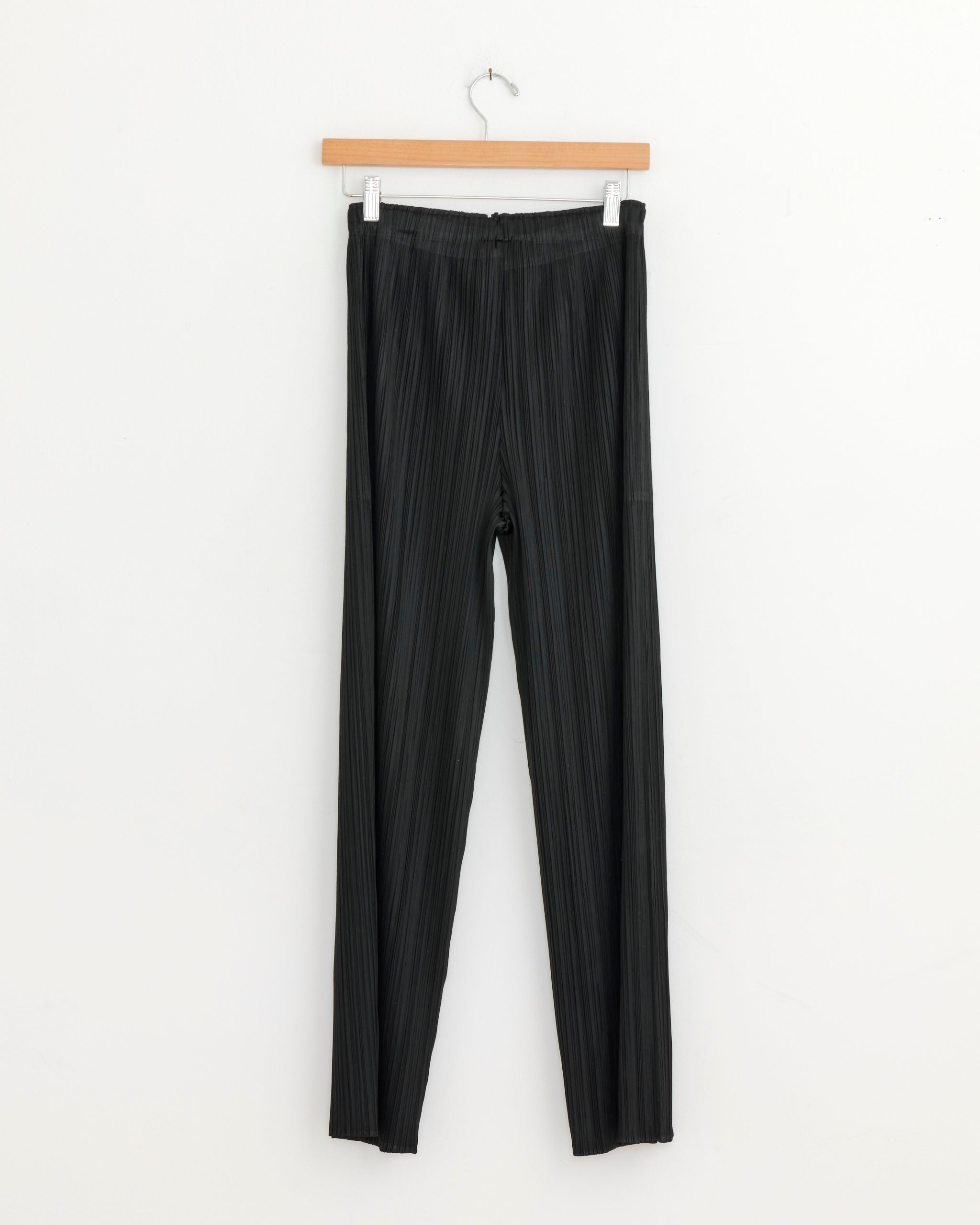 The MC Feb Pant in Black by Pleats Please Issey Miyake hangs on a wooden hanger against a plain white wall. Made in Japan, this is the Monthly Colors February Pant.
