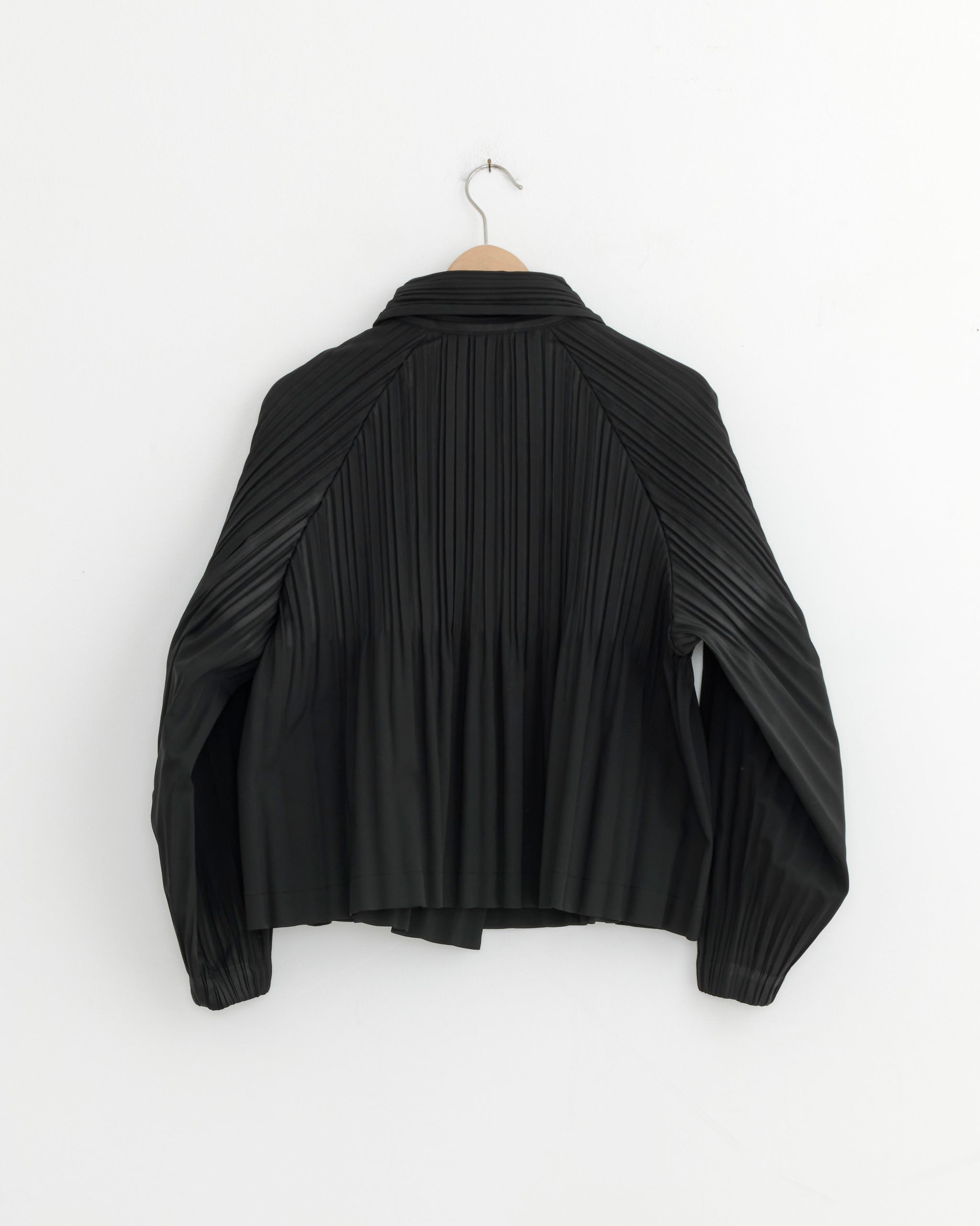 The Smooth Coat in Black by Pleats Please Issey Miyake, featuring long sleeves, a collar, and signature pleats, hangs on a wooden hanger against a white wall.