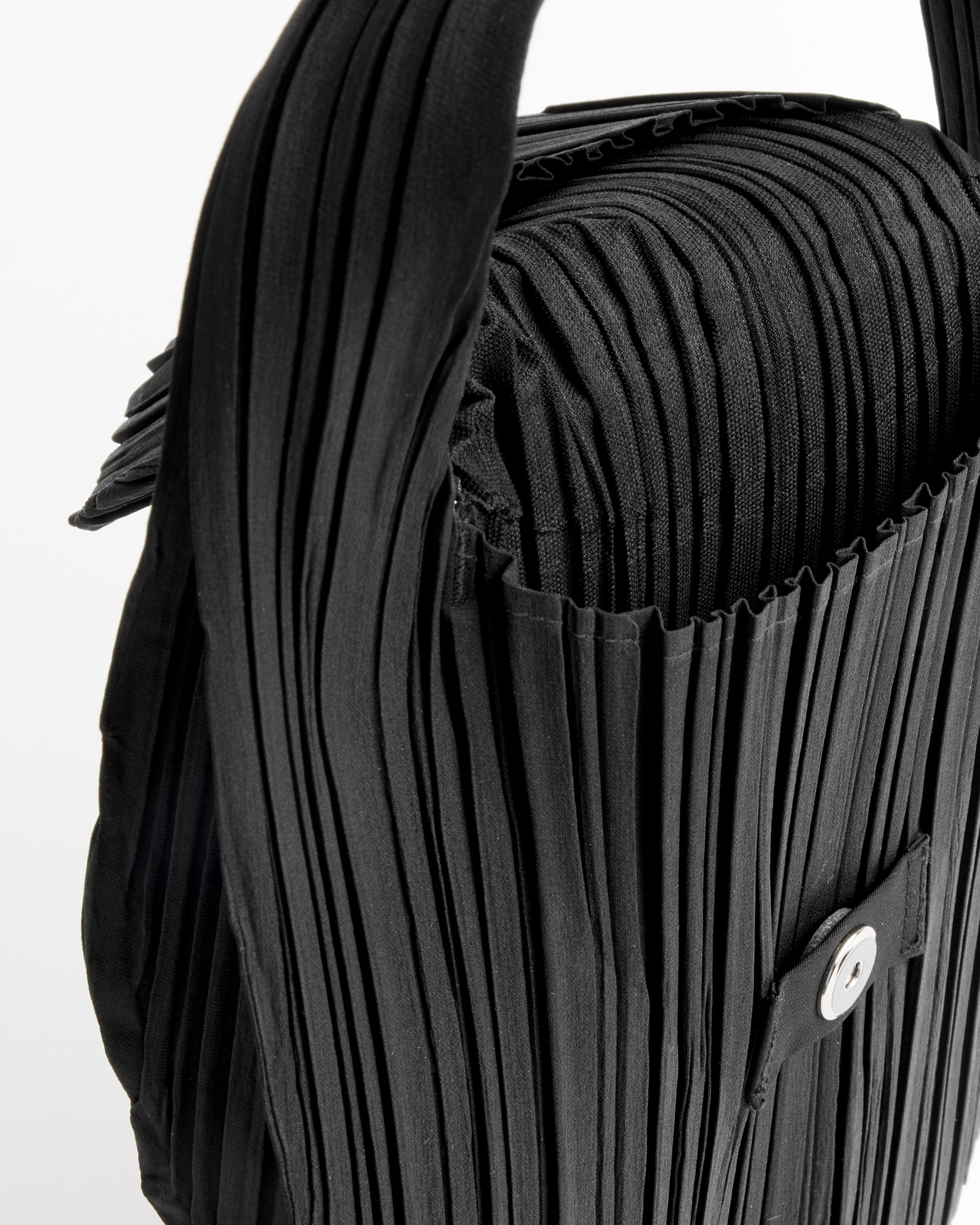 Close-up of the Pleats Please Issey Miyake Pleats Seeds Bag in Black, showing its ribbed pleated texture, handle, and main compartment with flap and button closure against a white background. Made in Japan.