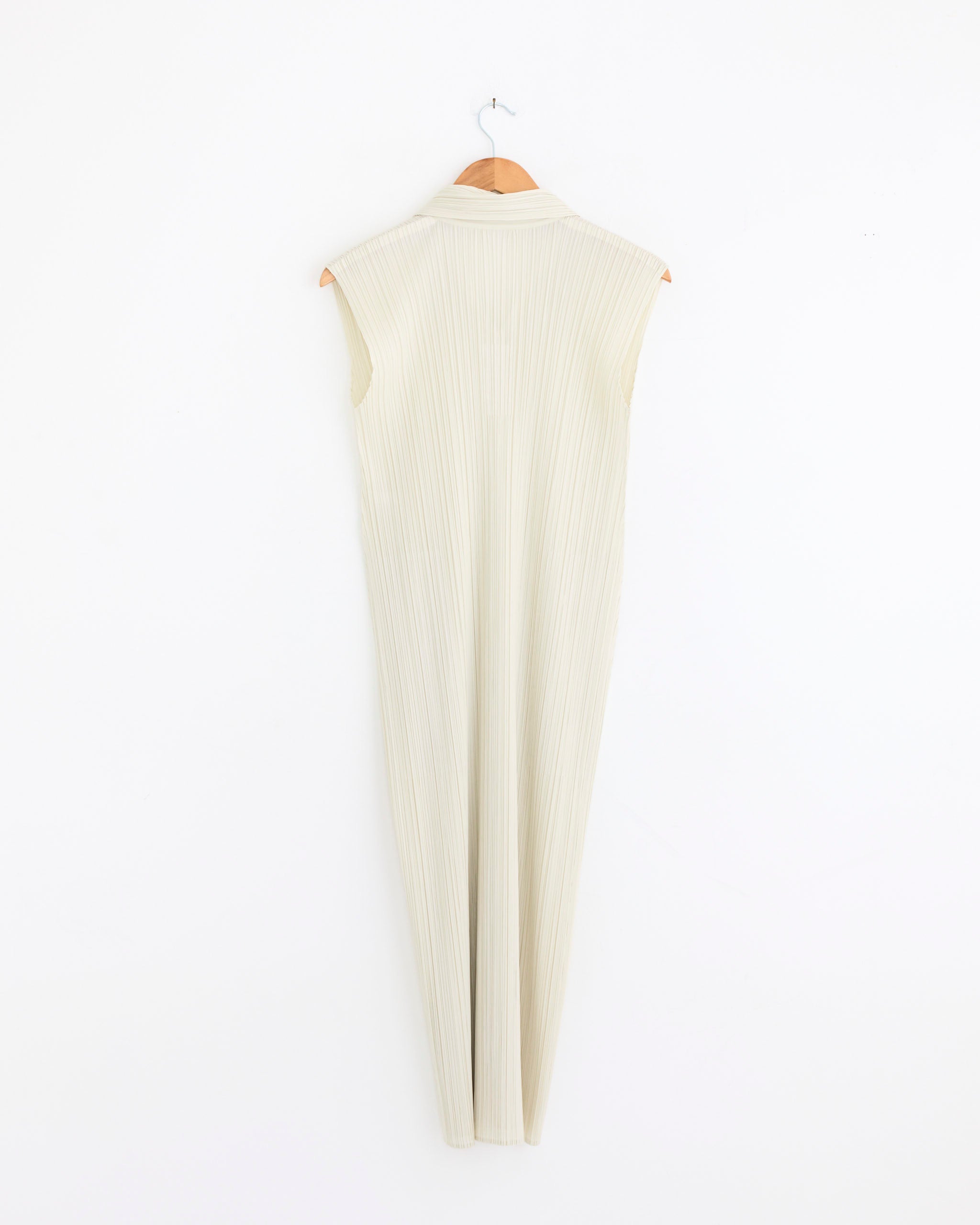 MC May Dress in Ivory by Pleats Please Issey Miyake, sleeveless and pleated, hangs on a wooden hanger against a plain white wall, displayed from the back.