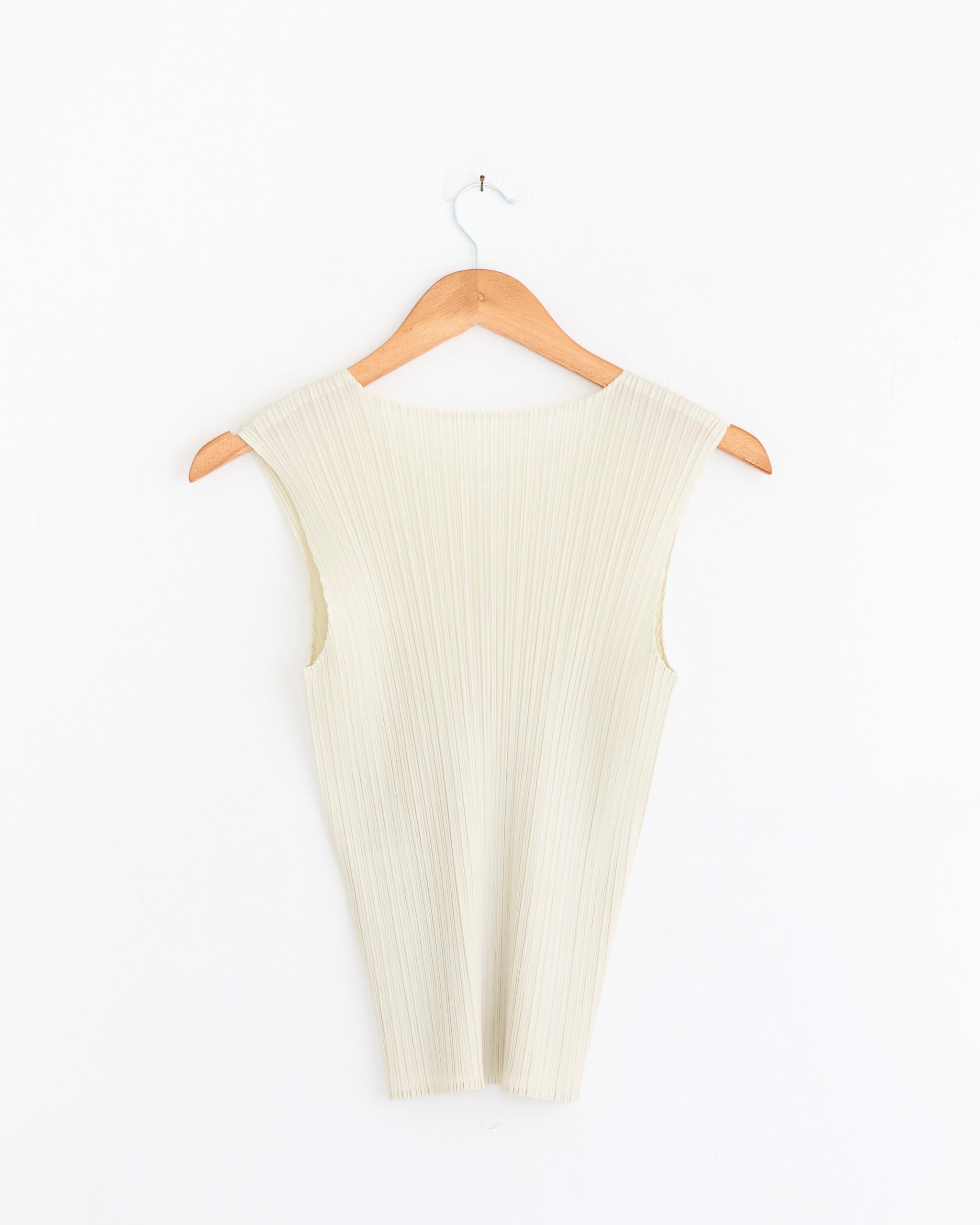 The Pleats Please Issey Miyake MC May Top in Ivory is a sleeveless, ribbed, pleated tank with a lightweight silhouette, shown hanging on a wooden hanger against a white background.
