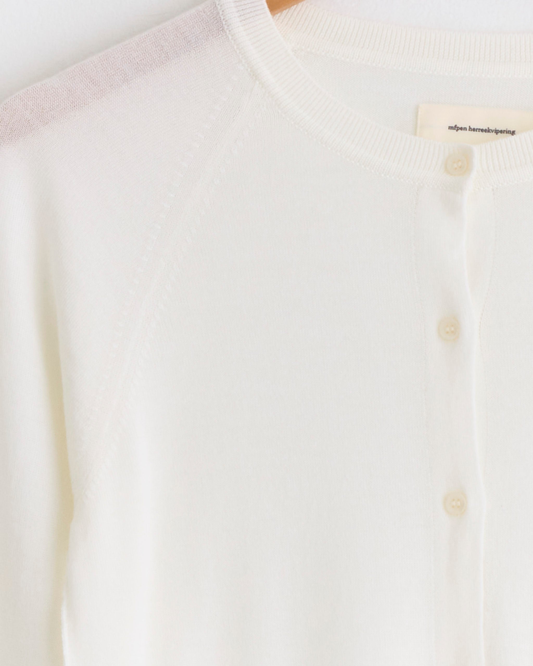 Close-up of the mfpen Appointment Cardigan in Ecru, hanging on a wooden hanger, with detailed view of buttons, ribbed neckline, and soft merino silk knit fabric.