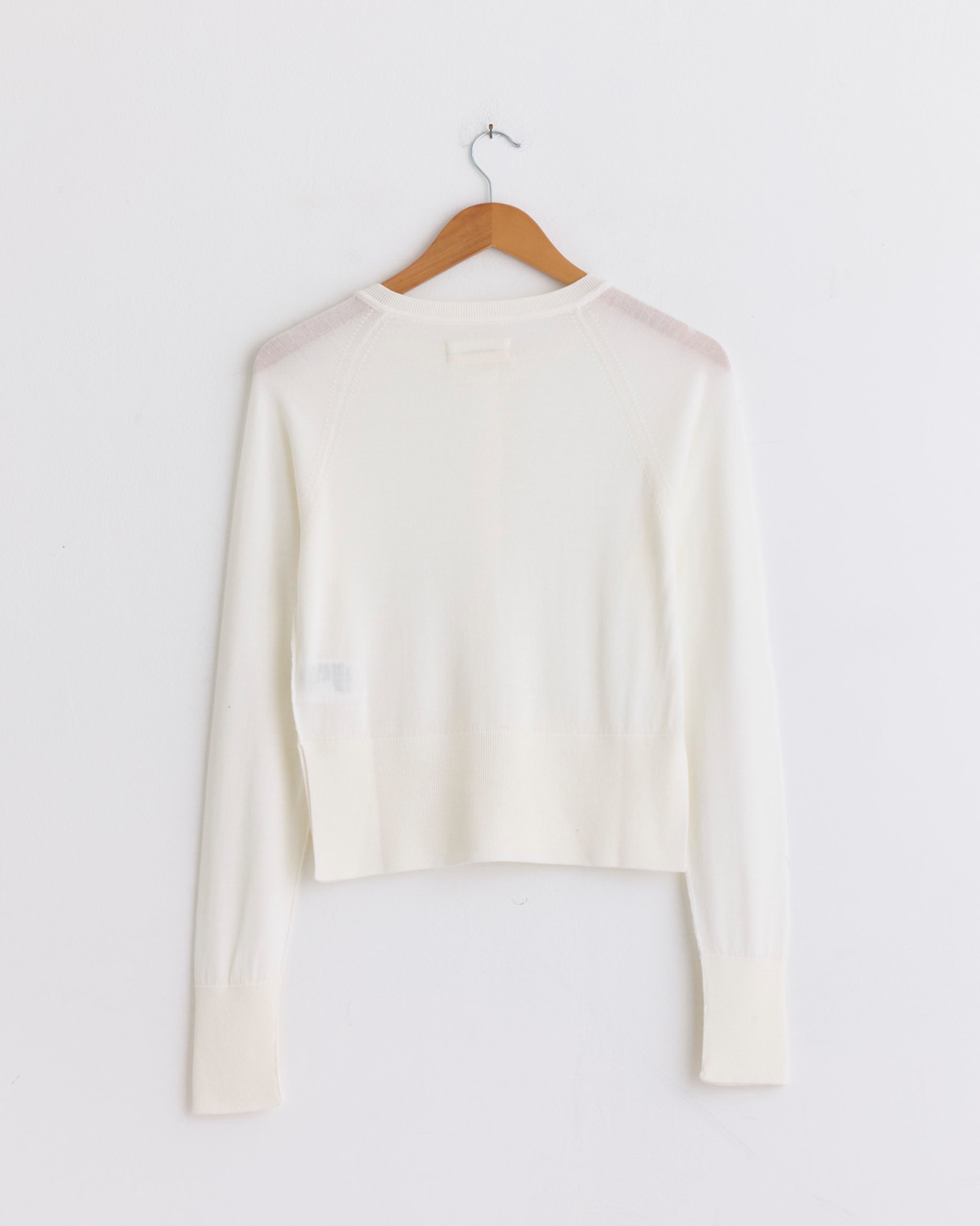 The mfpen Appointment Cardigan in Ecru, crafted from a merino silk knit, is shown from the back on a wooden hanger against a plain white wall.