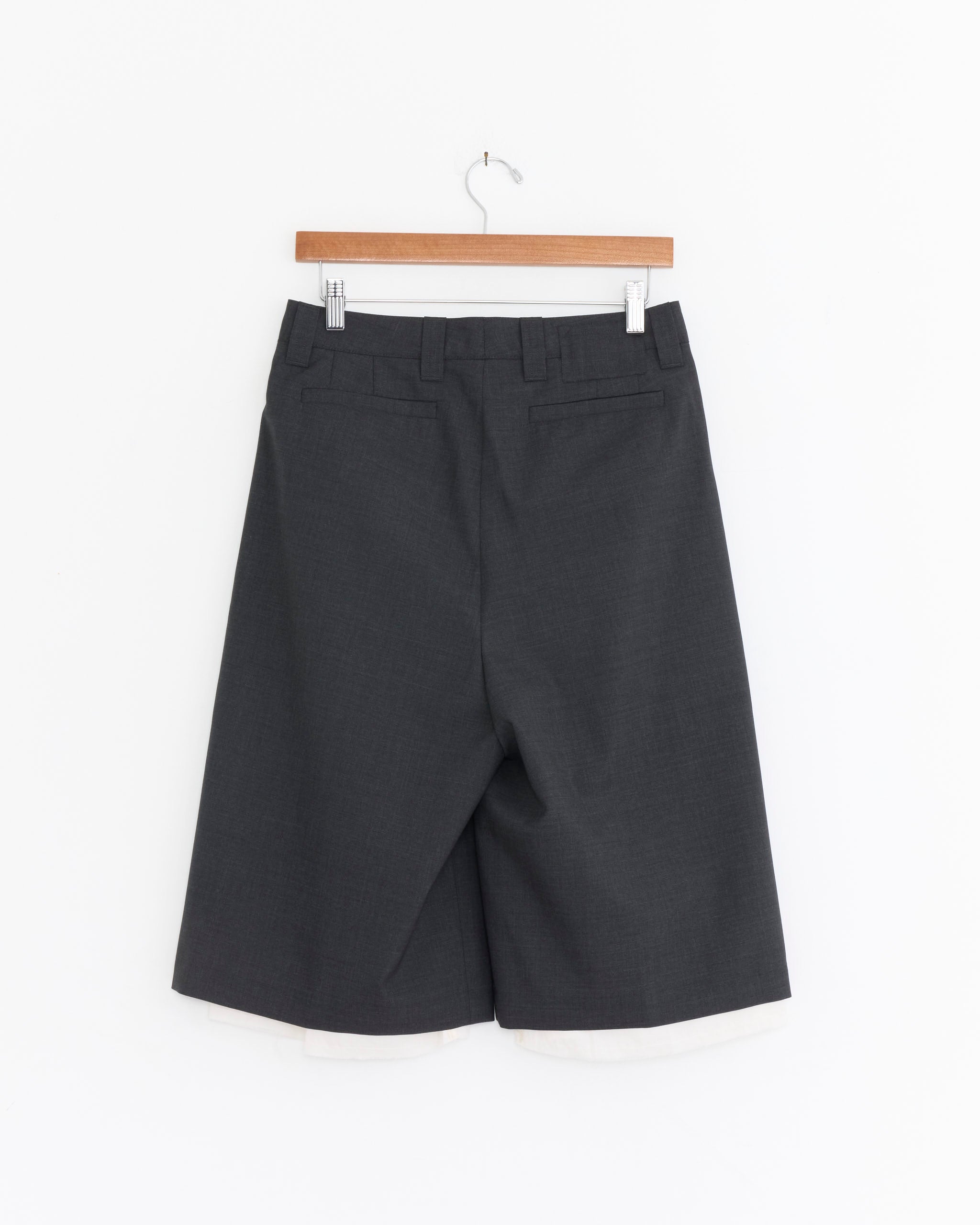 The Slash Short in Dark Grey by mfpen, crafted from tropical wool, features a wide straight fit with visible back pockets and belt loops. Shown hanging on a wooden hanger against a white background, displayed from the back.