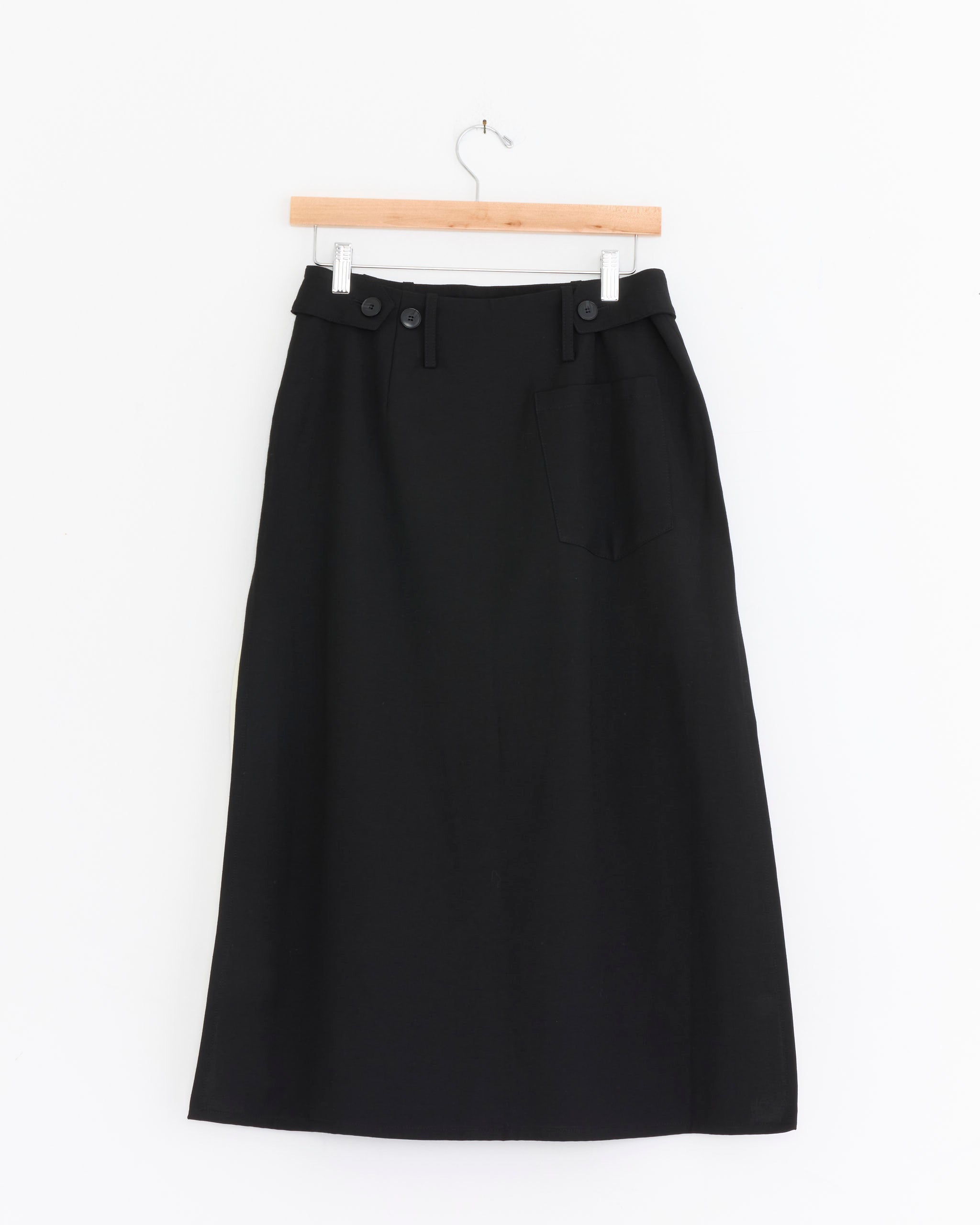 The mfpen Tab Skirt in Muted Black is a high-waisted, A-line cotton blend skirt featuring belt loops, button details at the waistband, and a single front pocket, shown on a wooden hanger against a white background.