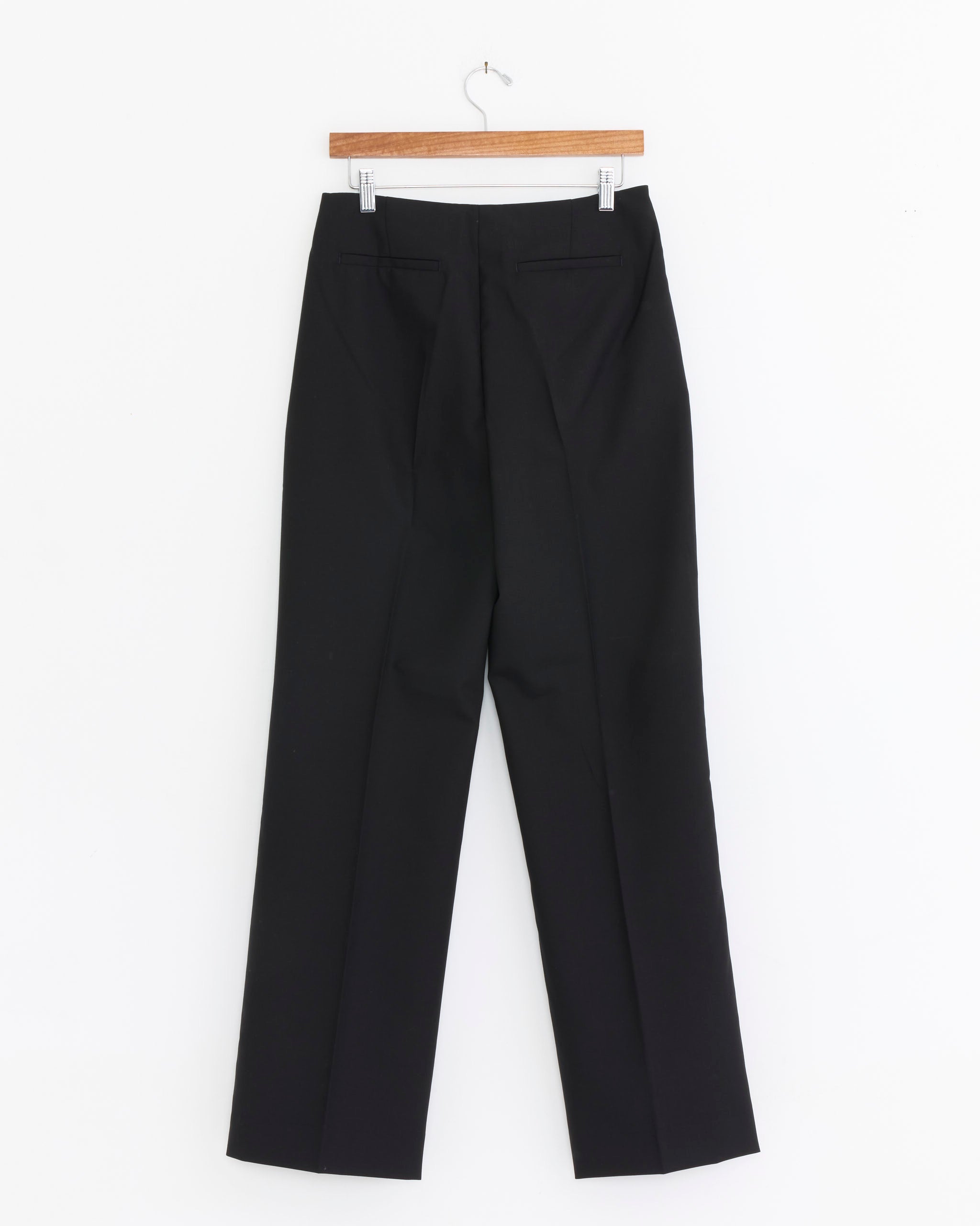 Mfpen Taper Trouser in Black, crafted from tropical wool, hangs on a wooden hanger against a white background. Shown from the back, it features straight fit legs and two welt pockets.
