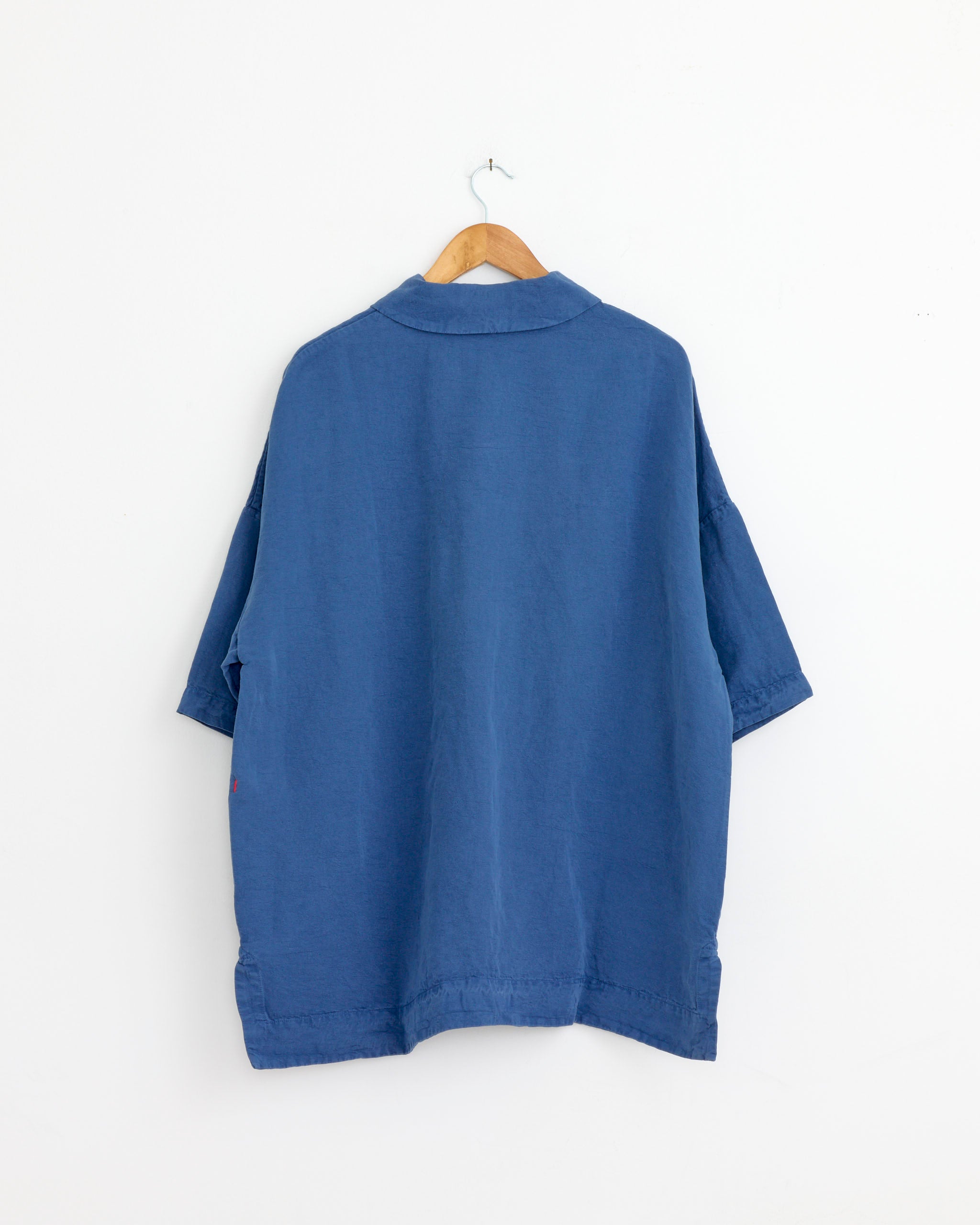 The Casey Casey Equerre Polo in Blue, a loose-fitting, short-sleeve polo made from heavy jersey, is displayed from the back on a wooden hanger against a plain white wall.