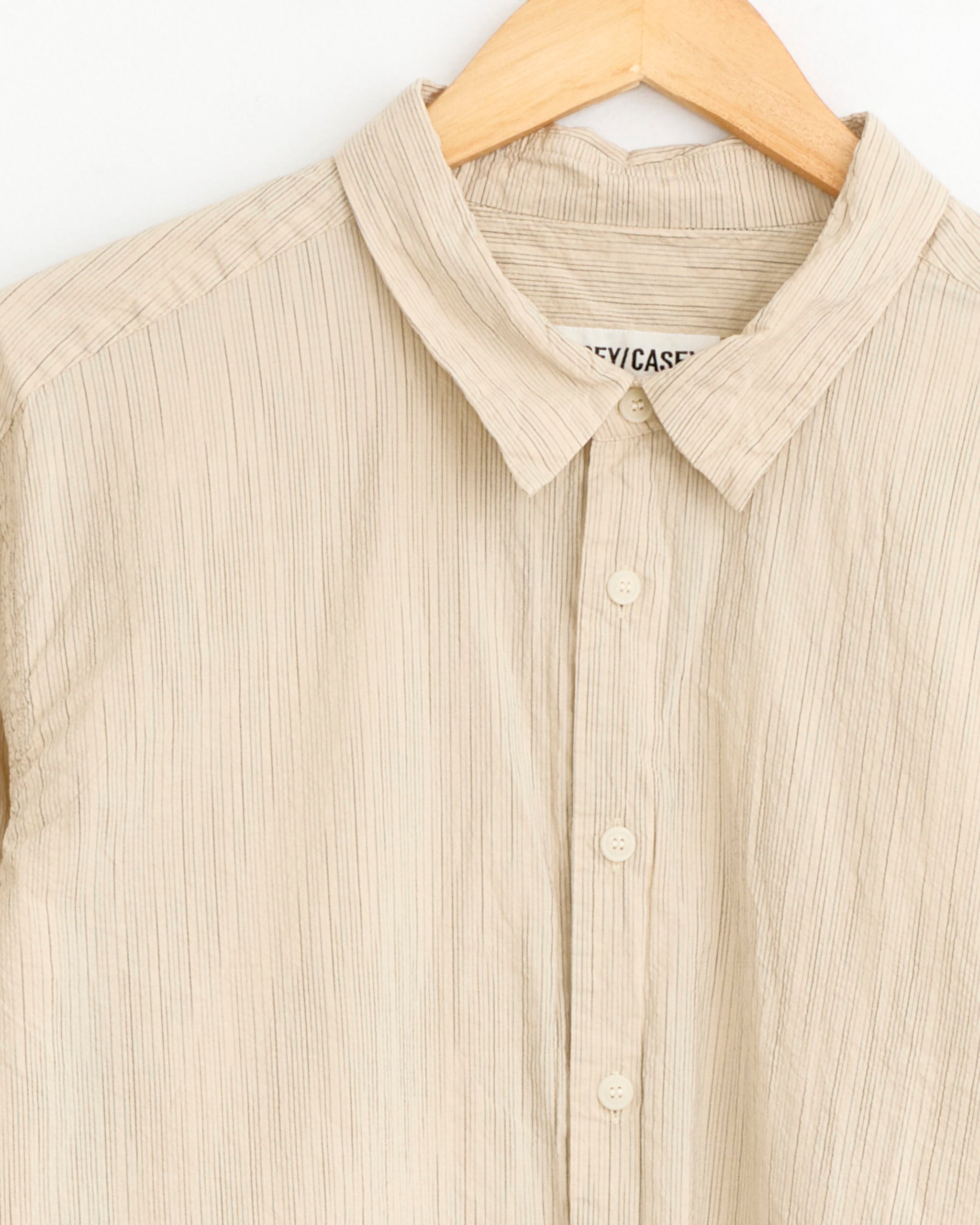 A close-up of the Casey Casey Fabiano Shirt in Nat Stripe, a beige, long-sleeve cotton shirt with subtle vertical stripes, shown on a wooden hanger against a white background.