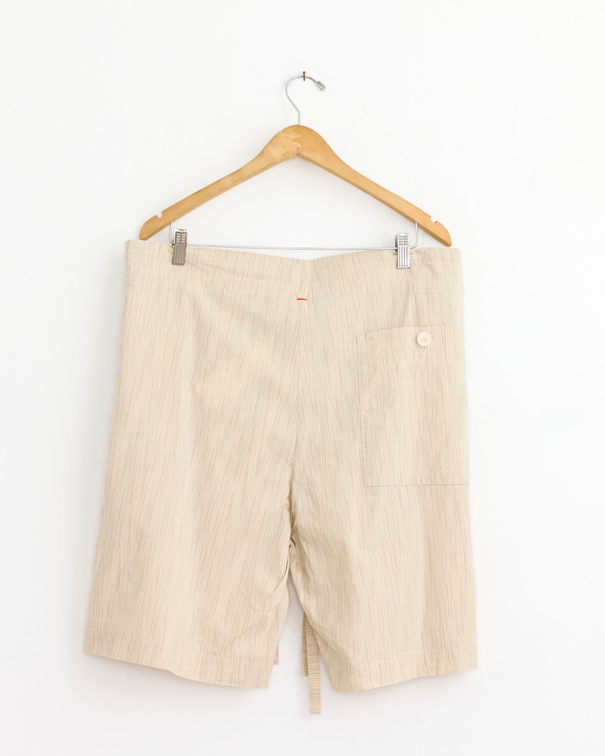 The Denmark Short in Nat Stripe by Casey Casey hangs on a wooden hanger against a white wall. These wide-leg cotton shorts feature a subtle vertical stripe, back pocket with button closure, and a lightweight, casual style.