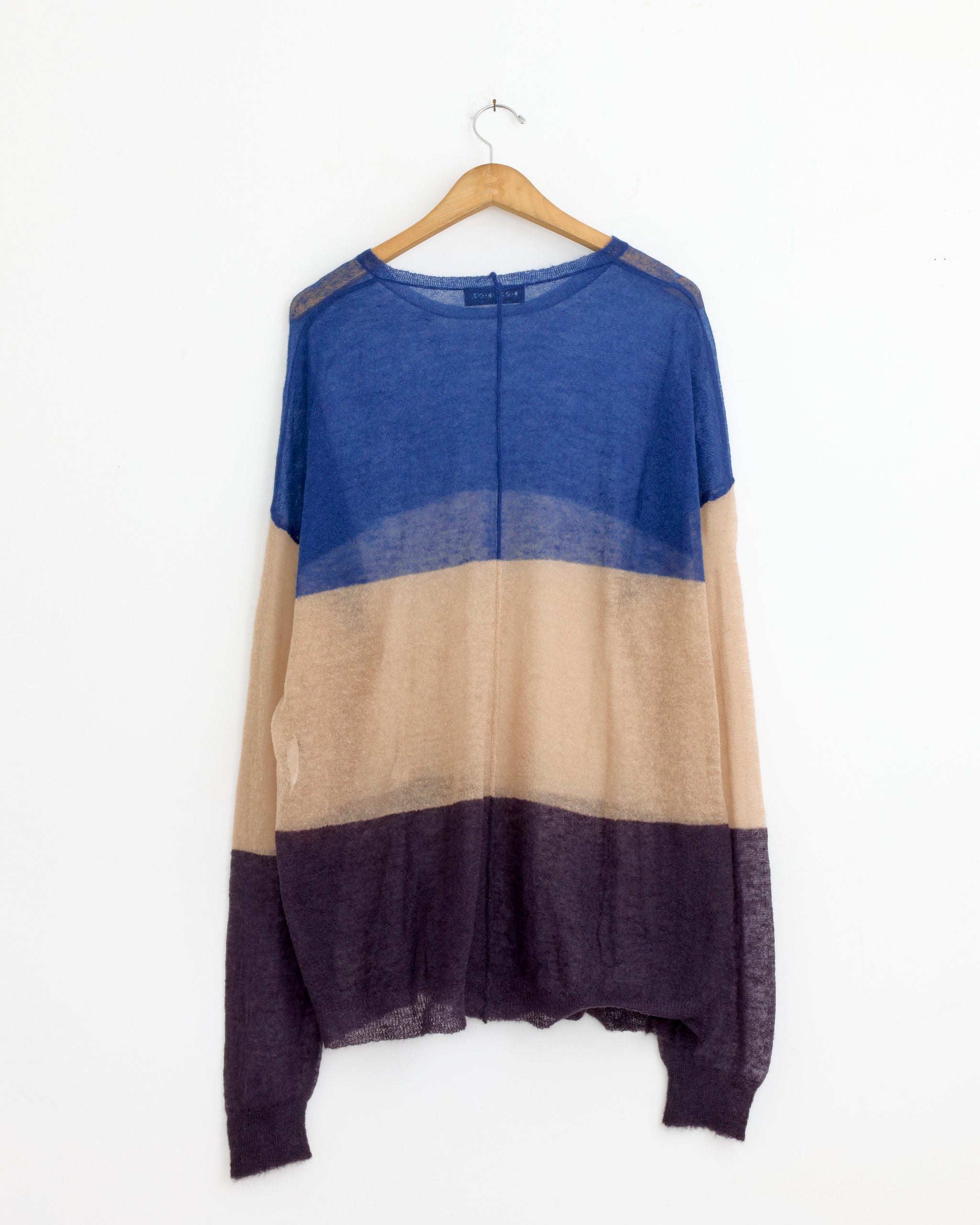 The Casey Casey Jumper in Stripes, a loose, long-sleeved oversized top with blue, beige, and dark purple horizontal blocks, hangs on a wooden hanger against a white wall.
