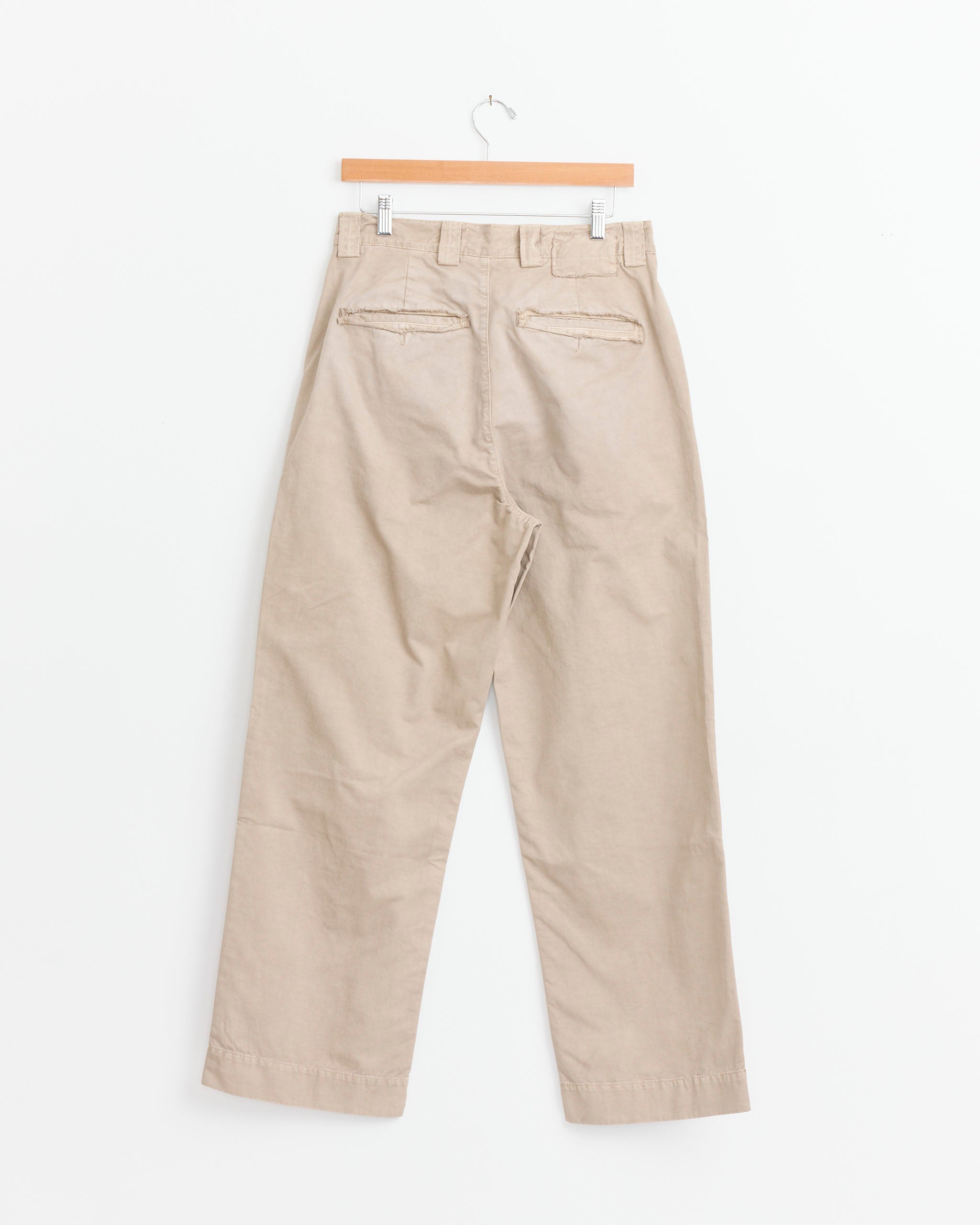 The mfpen Commission Chino in Stonewashed Sand, made from garment washed cotton twill, hangs on a wooden hanger against a white background, shown from the back with belt loops and two back welt pockets visible.