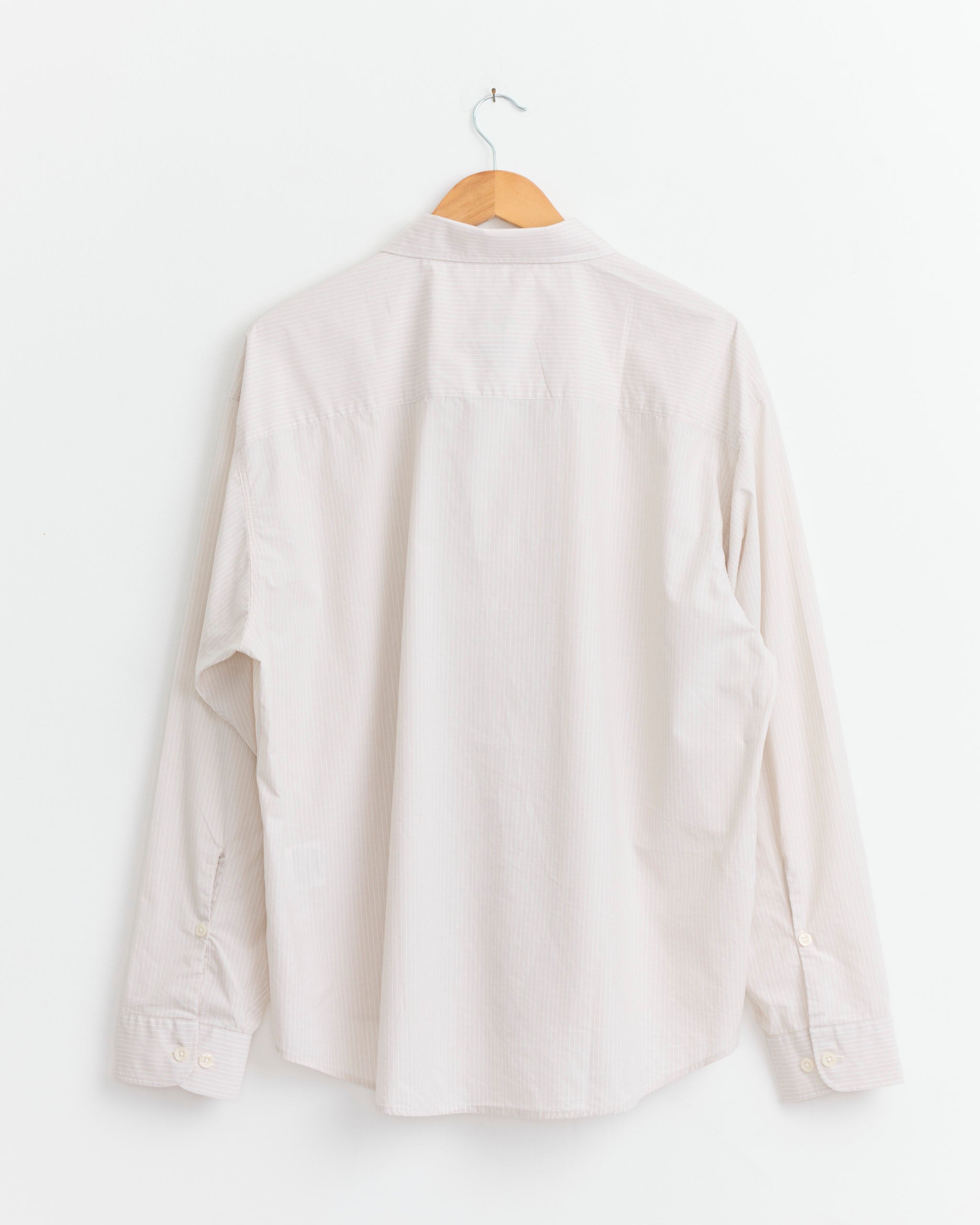 The mfpen Exact Shirt in Pale Office Stripe, featuring a light hue and long sleeves with a boxy fit, is shown hanging on a wooden hanger against a white wall, displayed from the back.