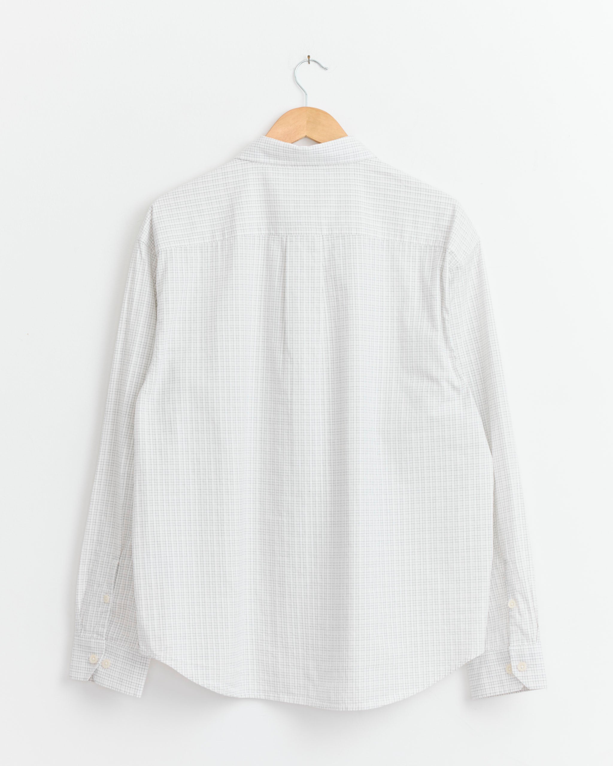 The mfpen Generous Shirt in White Connection Check hangs on a wooden hanger against a plain white wall, displayed from the back with its relaxed fit and buttoned cuffs visible.