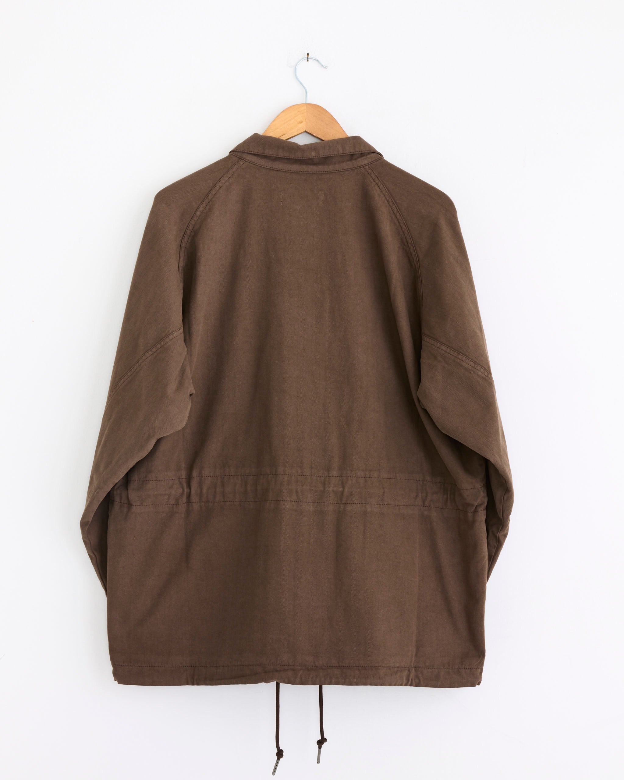 The mfpen Terrain Jacket in Brown, shown from the back on a wooden hanger against a white wall, features a relaxed fit, long sleeves, a collar, and drawstrings at the bottom hem.