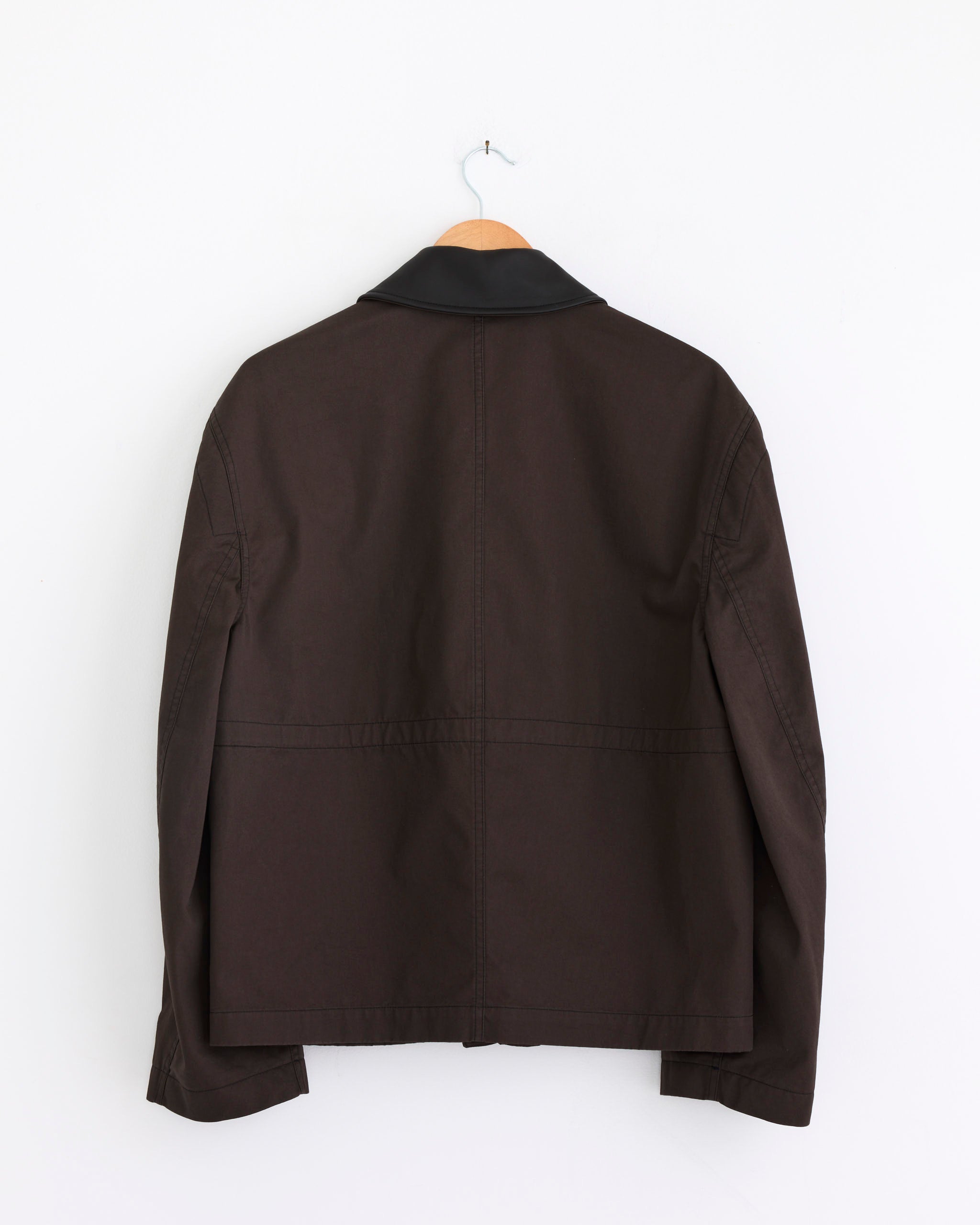 The Lemaire Leather Collar Blouson in Ristretto, a dark brown cotton twill jacket with a lamb leather collar, is shown from the back hanging on a wooden hanger against a plain white wall.