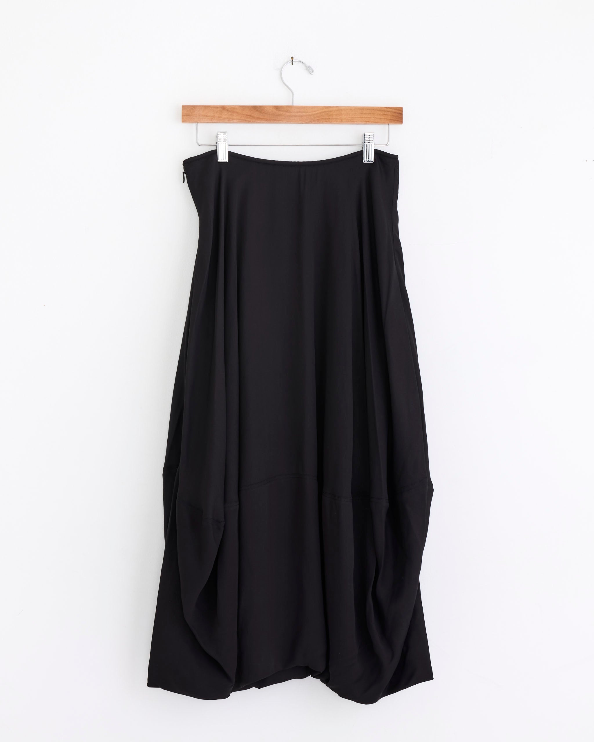 The Balloon Skirt in Granite by Lemaire is a long, loose-fitting black skirt with a draped asymmetrical hem and rounded silhouette in viscose crêpe de Chine, shown hanging on a wooden hanger against a plain white background.
