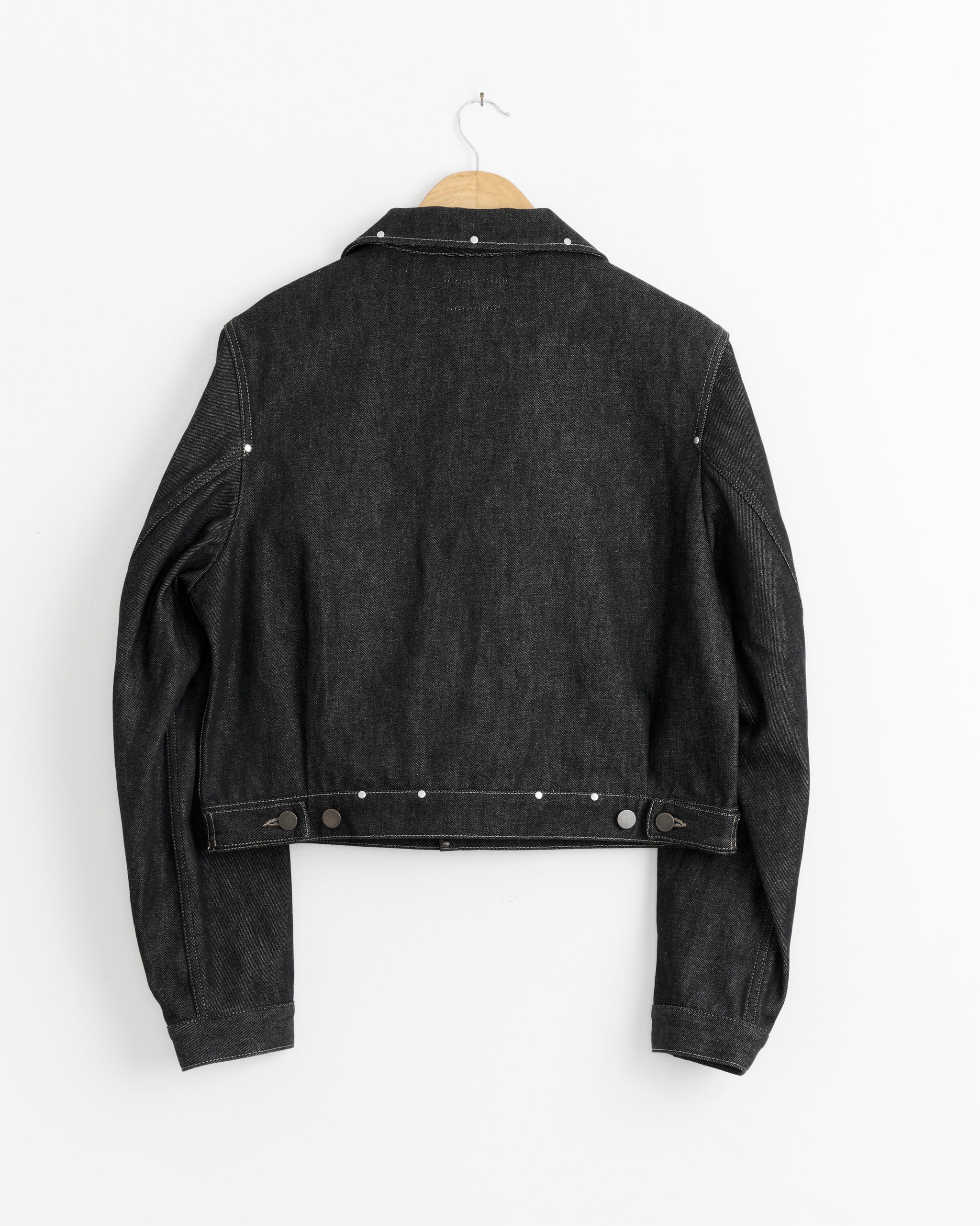 The Lemaire Cropped Blouson With Studs in Slate is displayed from the back on a wooden hanger against a white wall, featuring silver studs and visible stitching details.