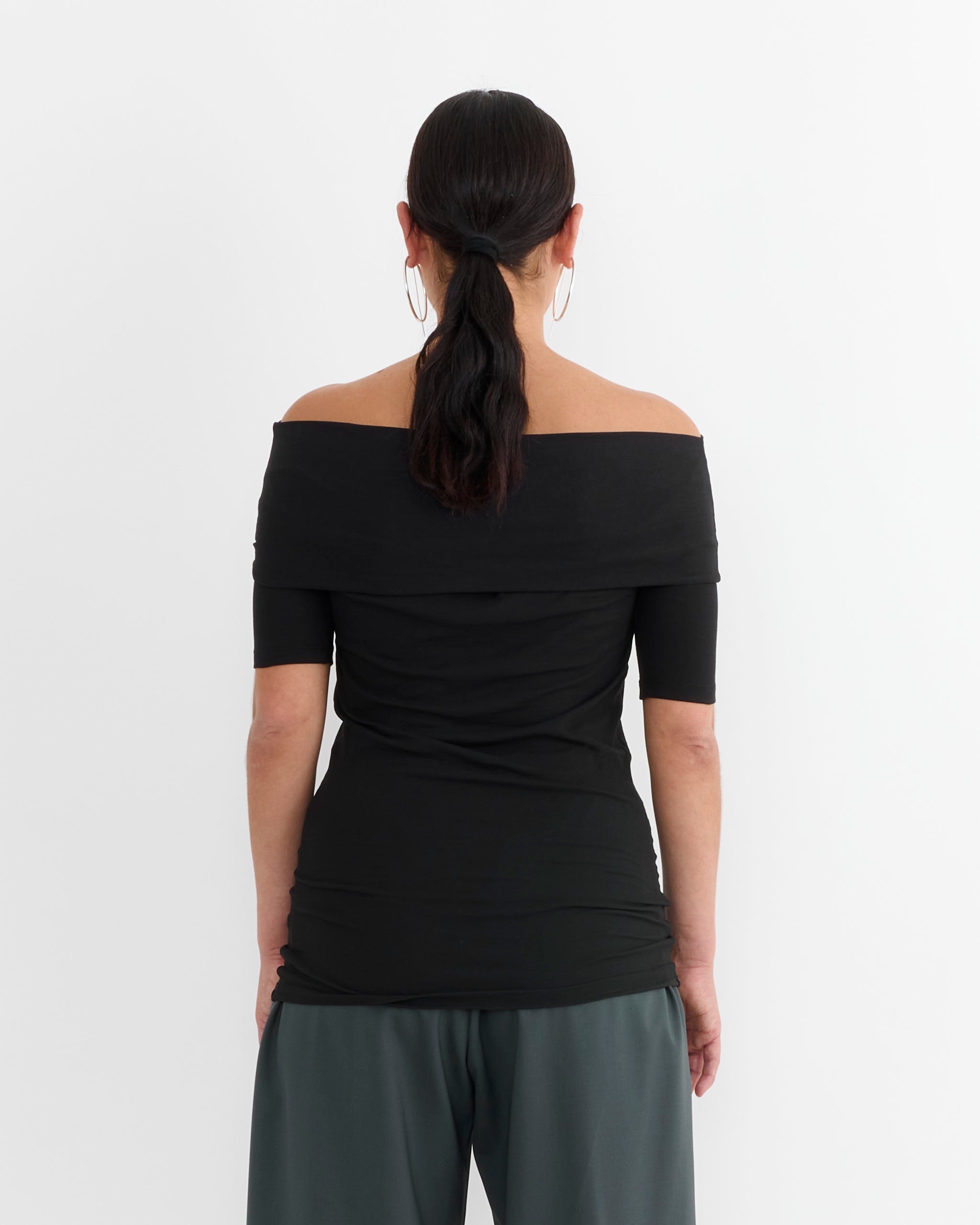 A woman with dark hair in a low ponytail is seen from behind, wearing the Lemaire Twisted Top in Black and dark green pants, standing against a plain white background.