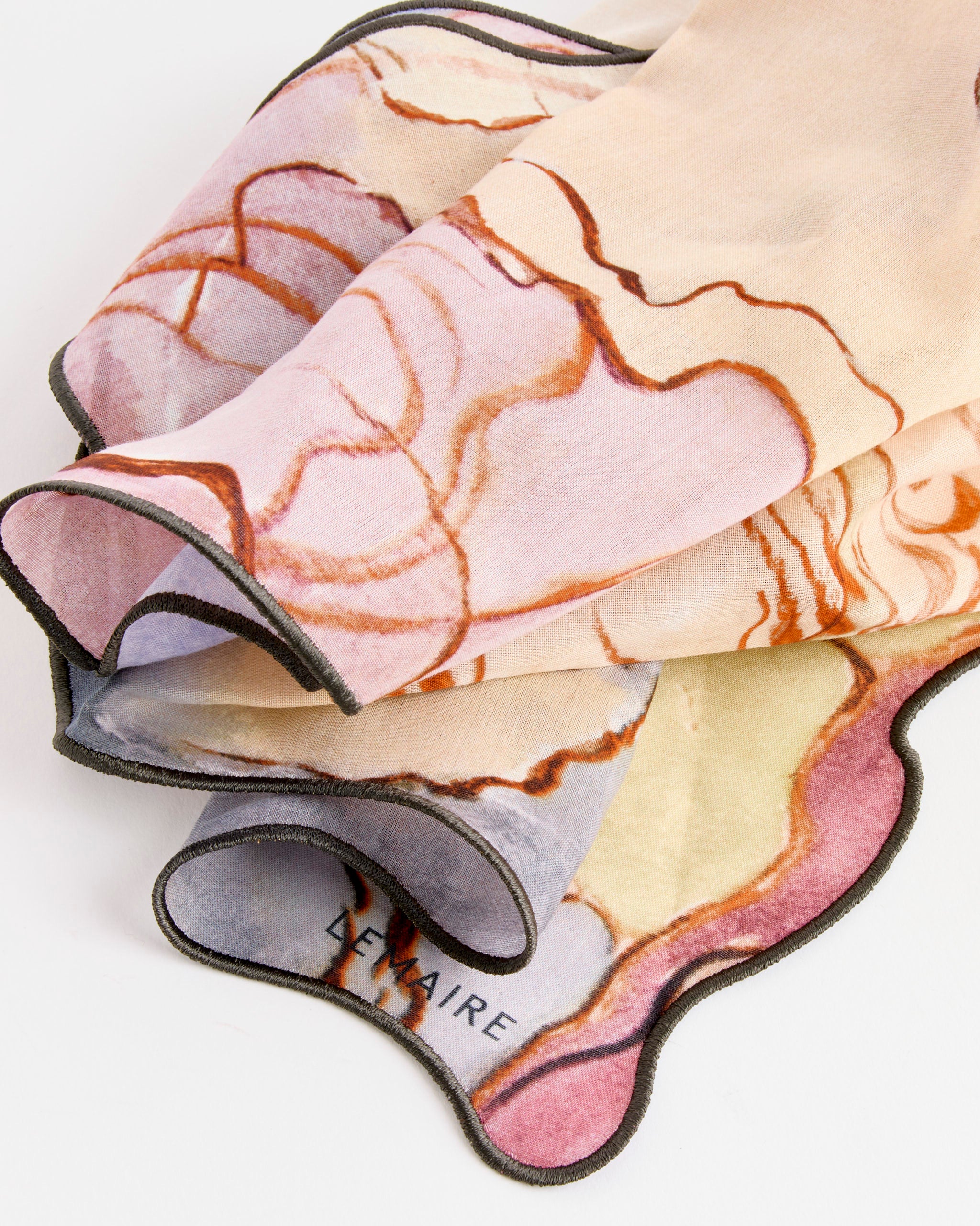 A close-up of Lemaire's Aquarel Irregulie Scarf Bandana in Multi shows wavy, marbled pastel patterns with dark edging and the LEMAIRE name printed in the corner, reminiscent of a watercolor artwork.