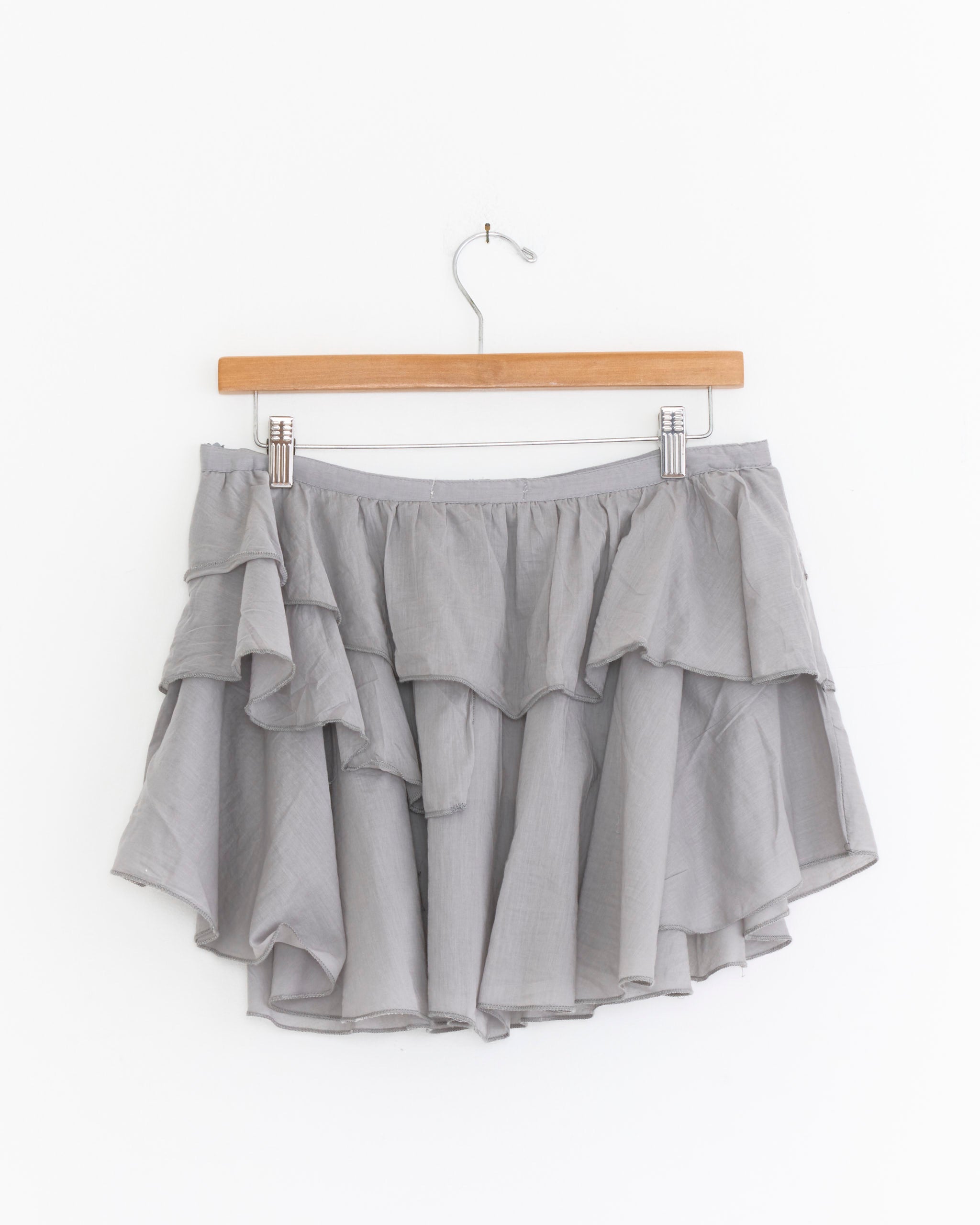 The Joan Mini Skirt in Grey by Gimaguas, featuring a light gray layered design with an asymmetrical hem, is displayed on a wooden hanger against a plain white background.