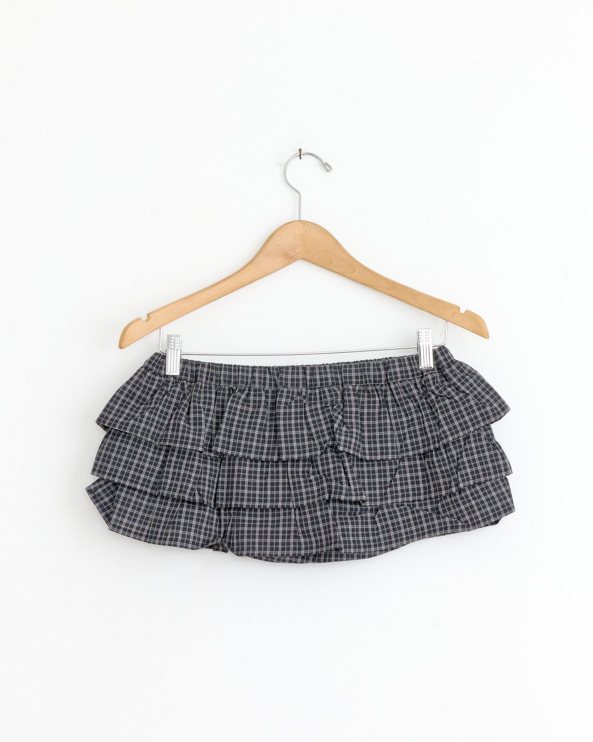 The Gimaguas Jardin Short in Black Check, a black-and-white plaid ruffle mini skirt, hangs on a wooden hanger against a plain white background.