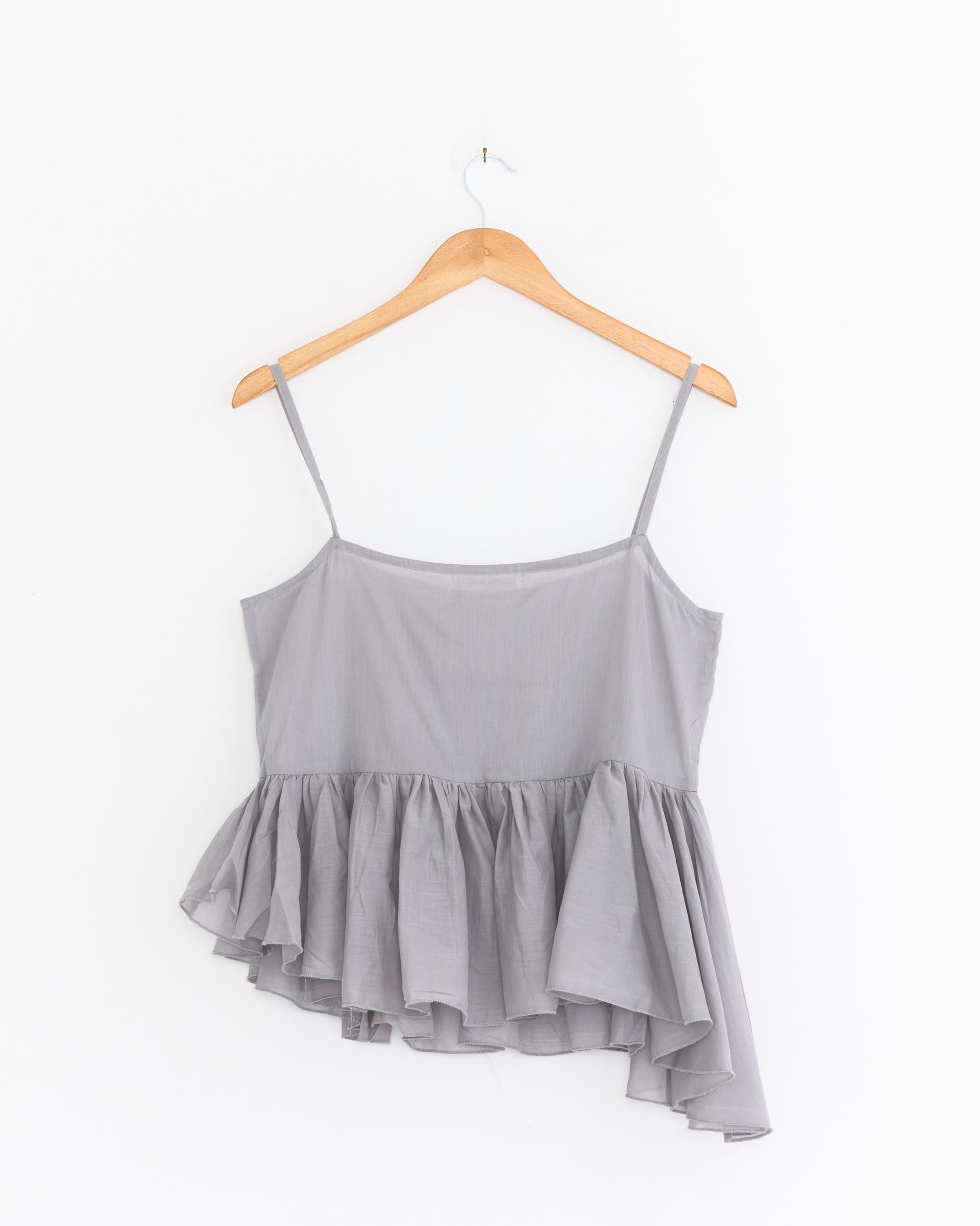 The Joan Top in Grey by Gimaguas, featuring thin straps and an asymmetric ruffled hem, hangs sleeveless on a wooden hanger against a plain white background.