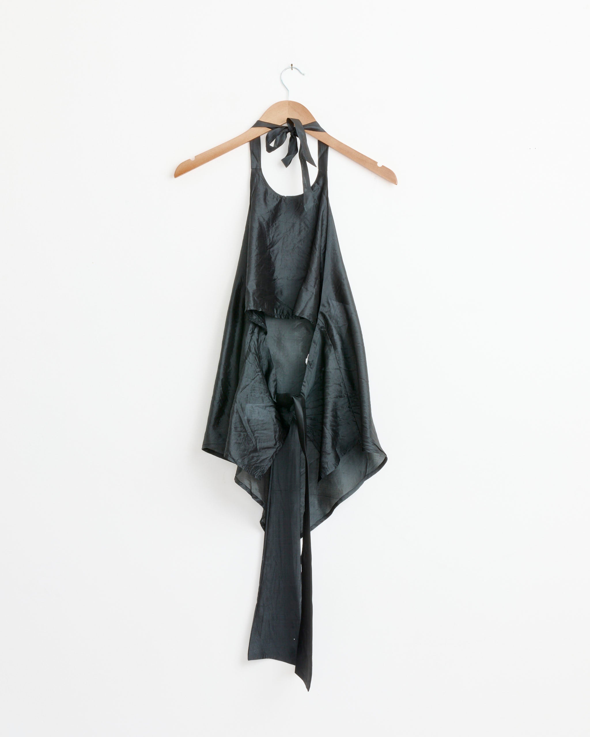 The Belle Top in Black by Gimaguas features silk fabric, a high neckline, sleeveless cut, adjustable lace-up back, and asymmetrical long ties. Shown on a wooden hanger against a plain white background.