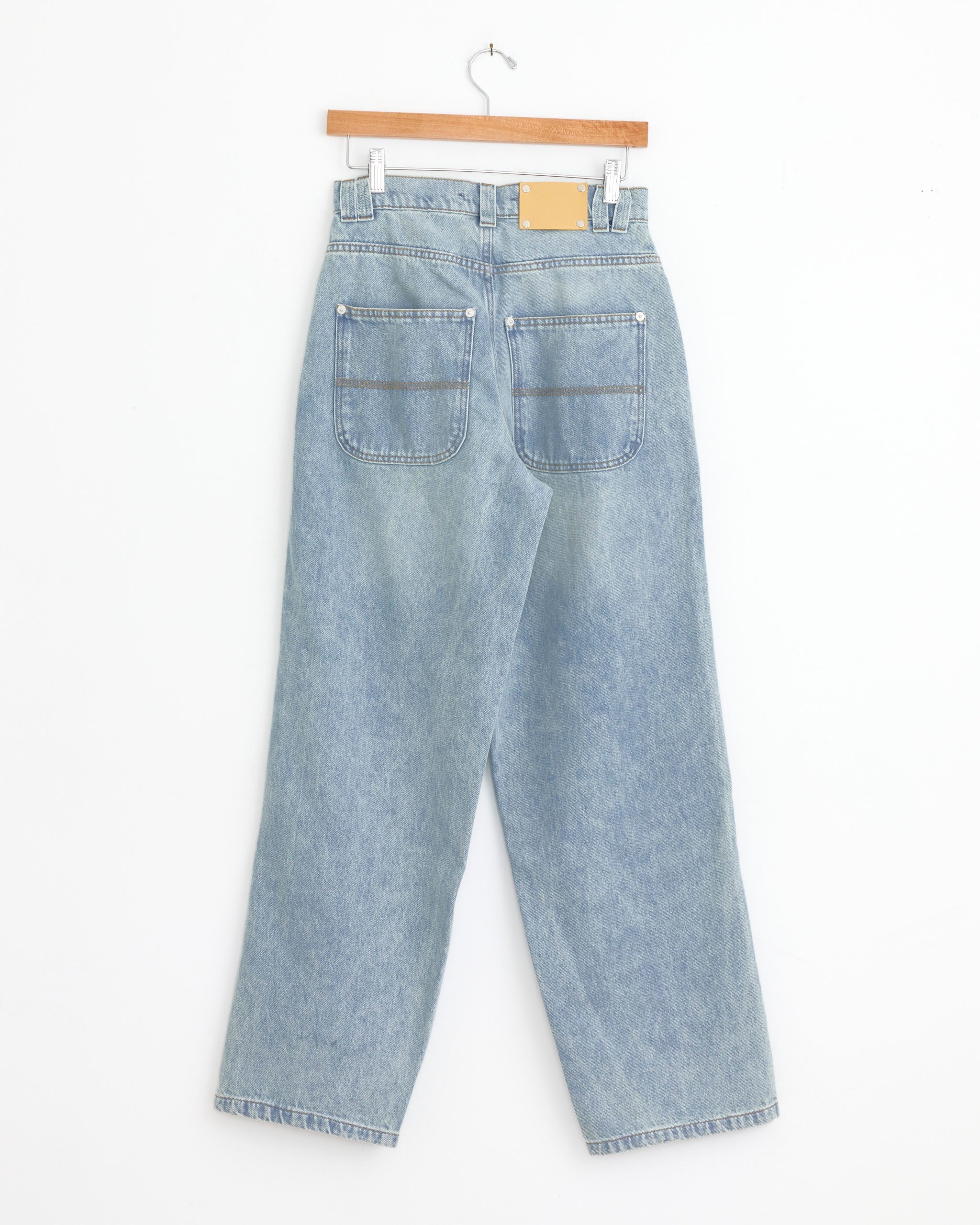 The Baggy Trouser in Washed Blue by Gimaguas hangs on a wooden hanger against a white wall, displayed from the back with two large rear pockets and a tan waistband patch.