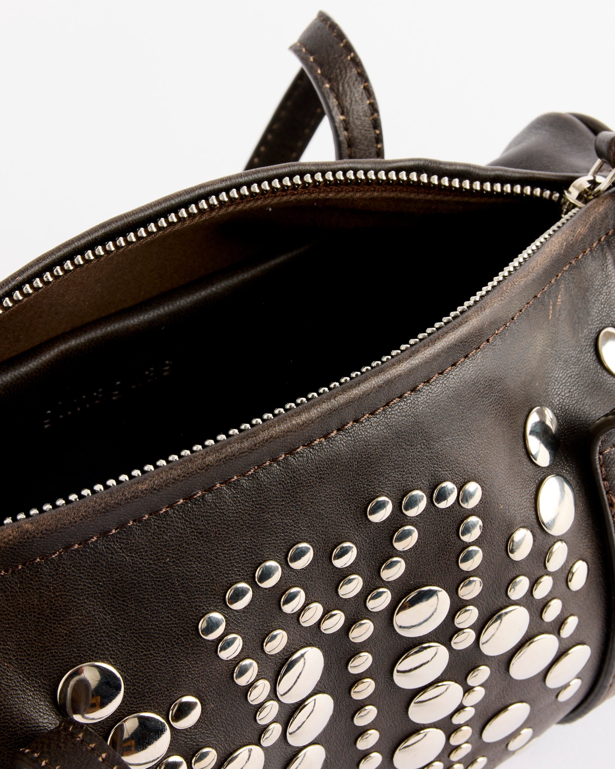 A close-up of the Gimaguas Sofia Faded Bag in Brown, made in Spain, featuring silver studs and a worn-in finish. The partially unzipped bag reveals its textured leather and intricate decorative details inside.
