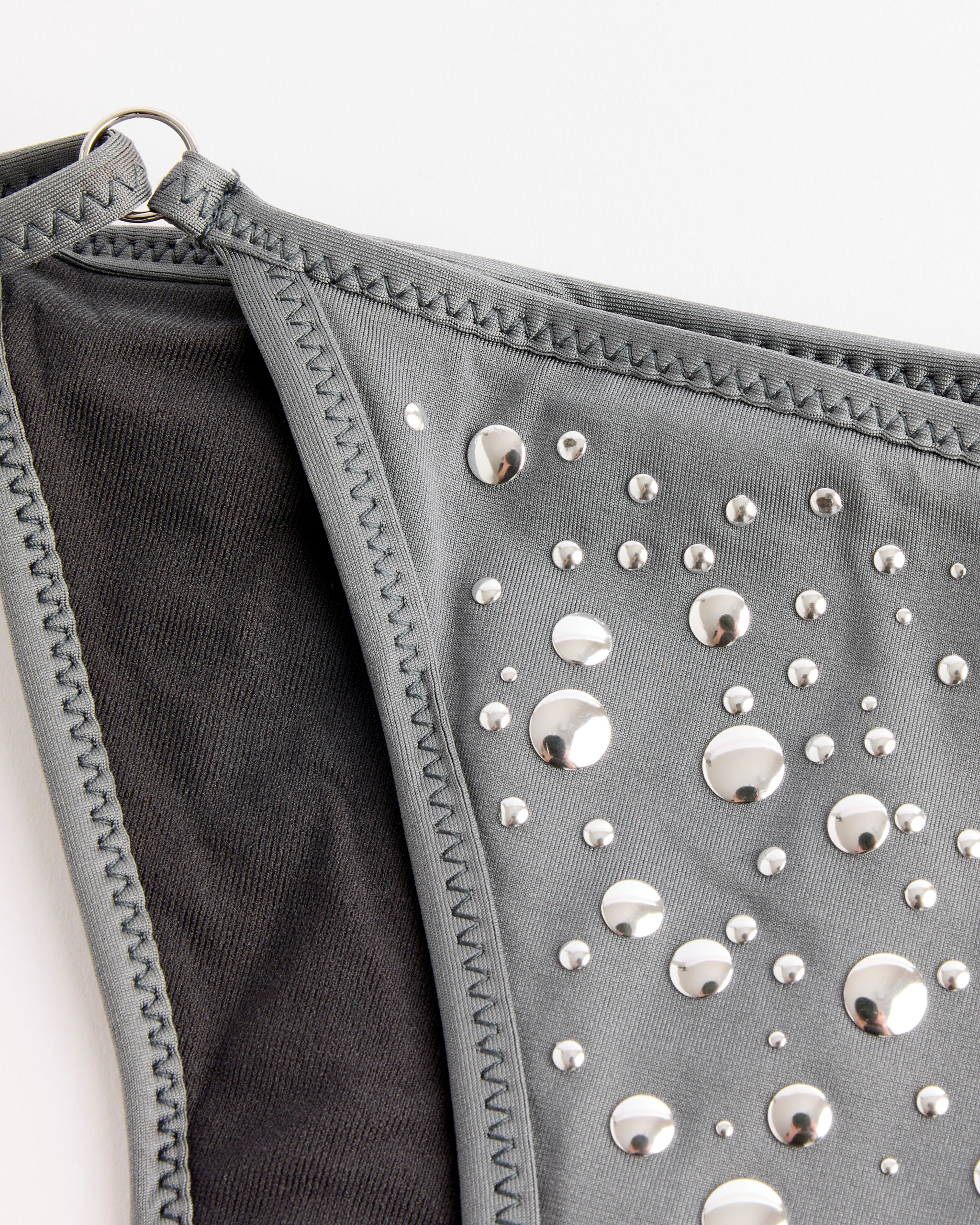 Close-up of the Gaia Bikini Bottom in Grey by Gimaguas, showing gray fabric with shiny metallic studs in different sizes, black lining, visible edge stitching, and a small metal ring at the corner.