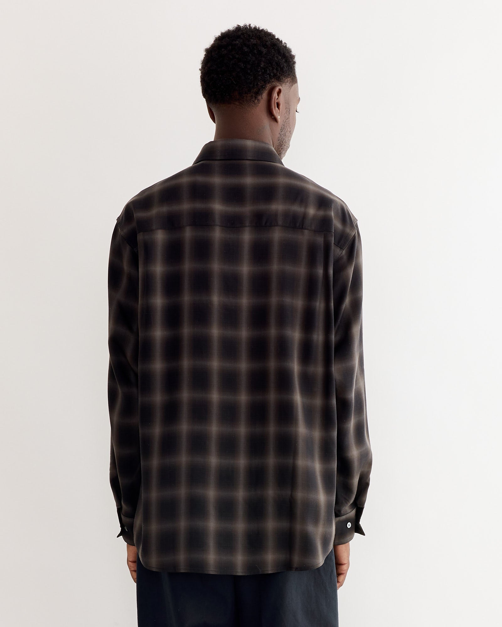 A man with short curly hair stands with his back to the camera, wearing the SMOCK Routine Shirt in Tencel Ombre Cacao and dark pants against a plain light background.