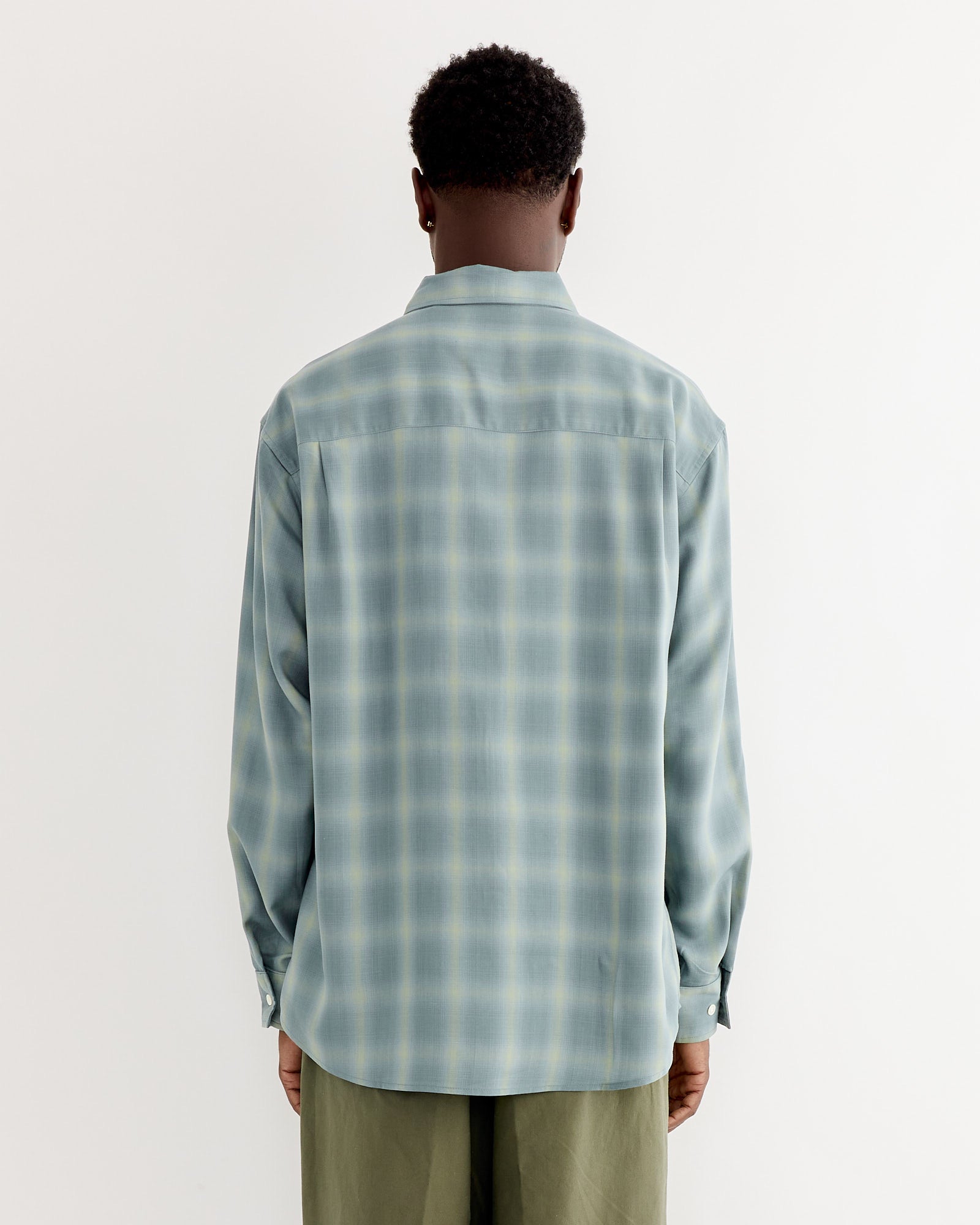 A person faces away from the camera in a SMOCK Routine Shirt in Tencel Ombre Azul and olive green pants, standing against a plain white background.