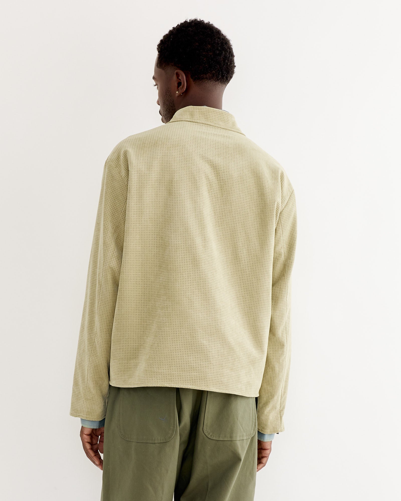 A man stands with his back to the camera, wearing the SMOCK Zip Jacket in Perforated Suede Sand and olive green pants against a plain light background.