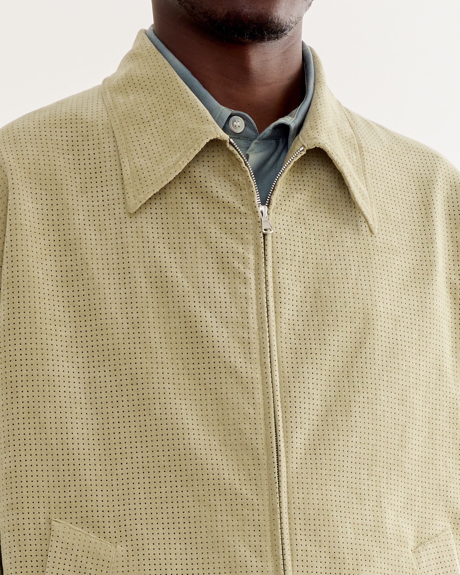 A close-up of a person wearing the SMOCK Zip Jacket in Perforated Suede Sand over a buttoned shirt. The light suede jacket features a textured, perforated pattern, with the person's face partially out of frame.