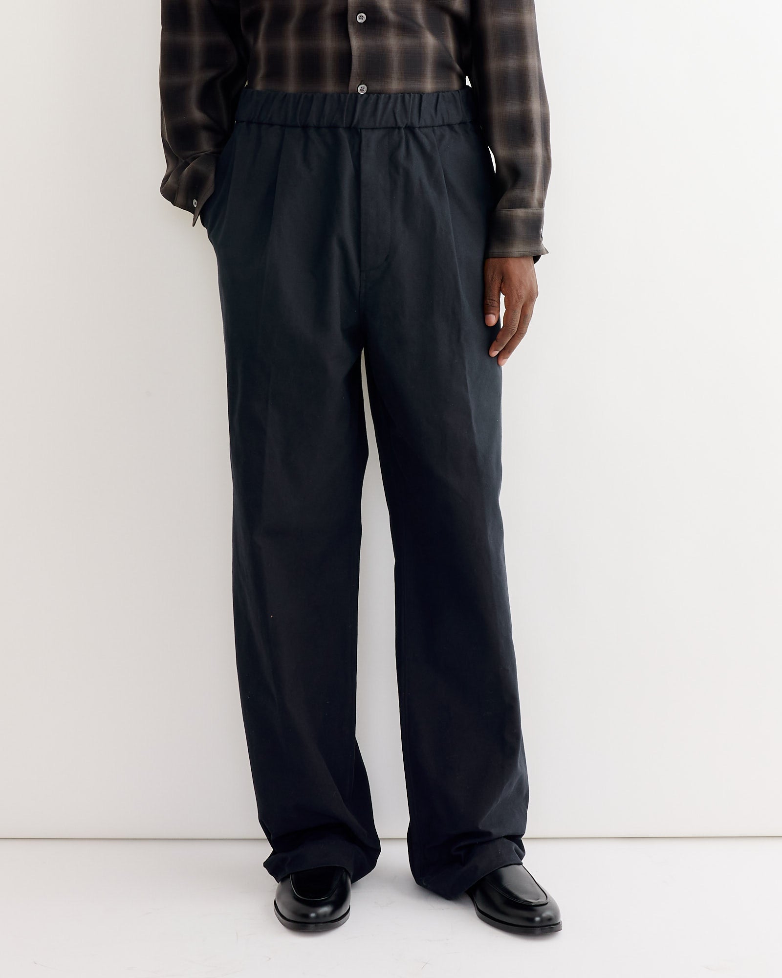 A person wearing SMOCK's Kogai Pant in Duck Canvas Black, black shoes, and a dark plaid shirt stands against a plain white background. The image is cropped from the shoulders down.