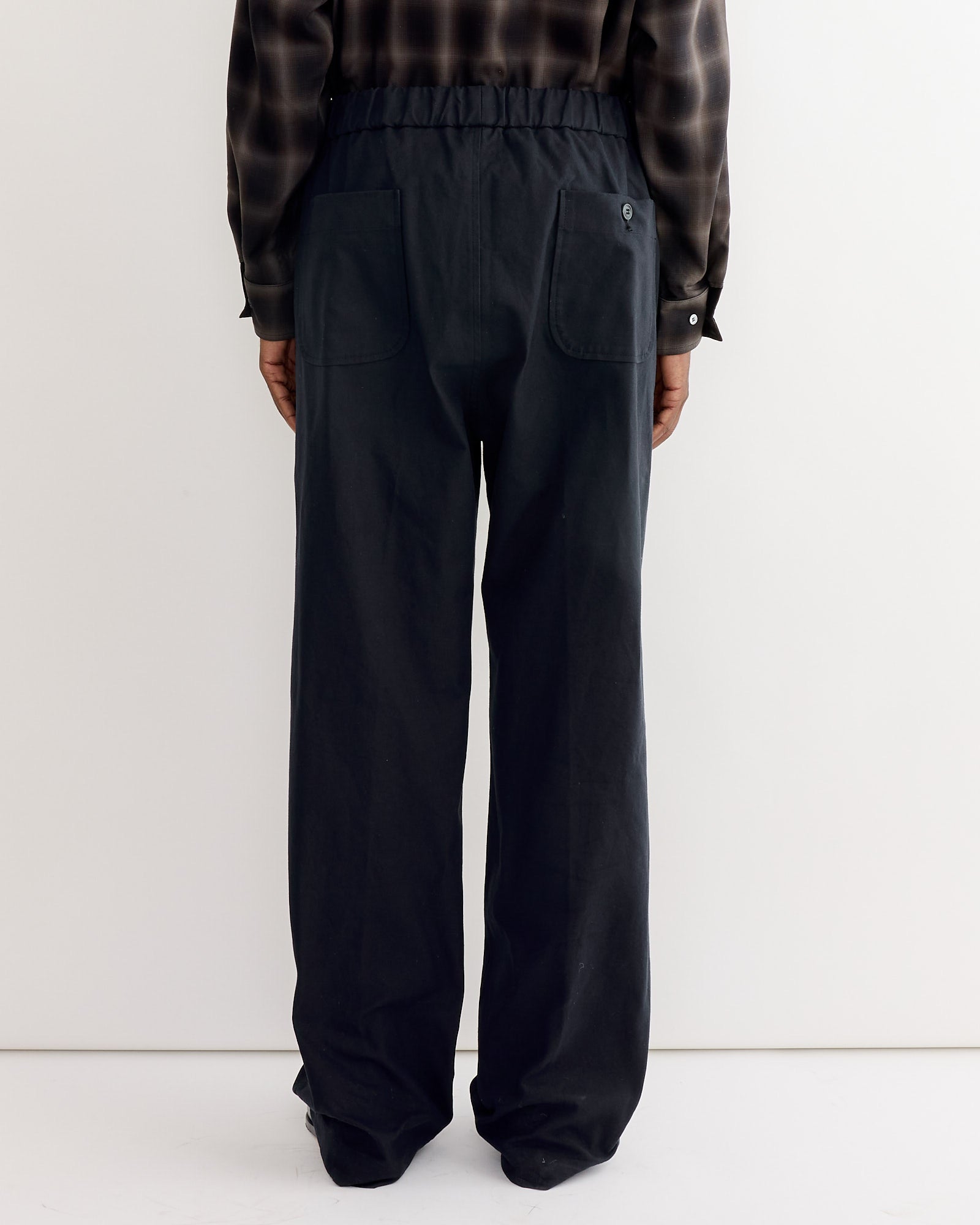 A person stands with their back to the camera wearing the SMOCK Kogai Pant in Duck Canvas Black and a dark plaid long-sleeve shirt, set against a plain white background.