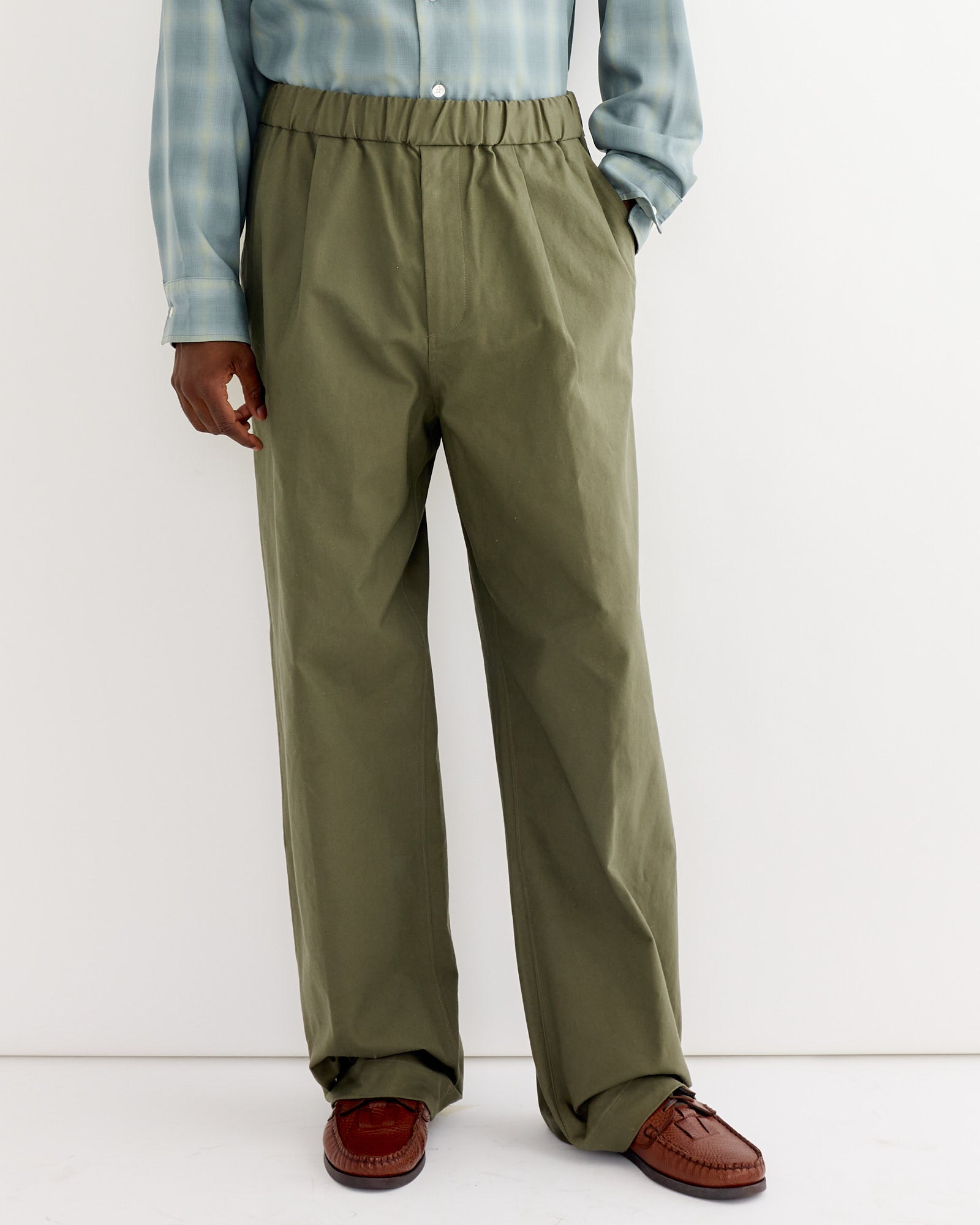 A person wears SMOCK’s Kogai Pant in Duck Canvas Olive with a light blue plaid shirt and brown loafers, sleeves rolled down, standing against a white background. The person’s face is not visible.