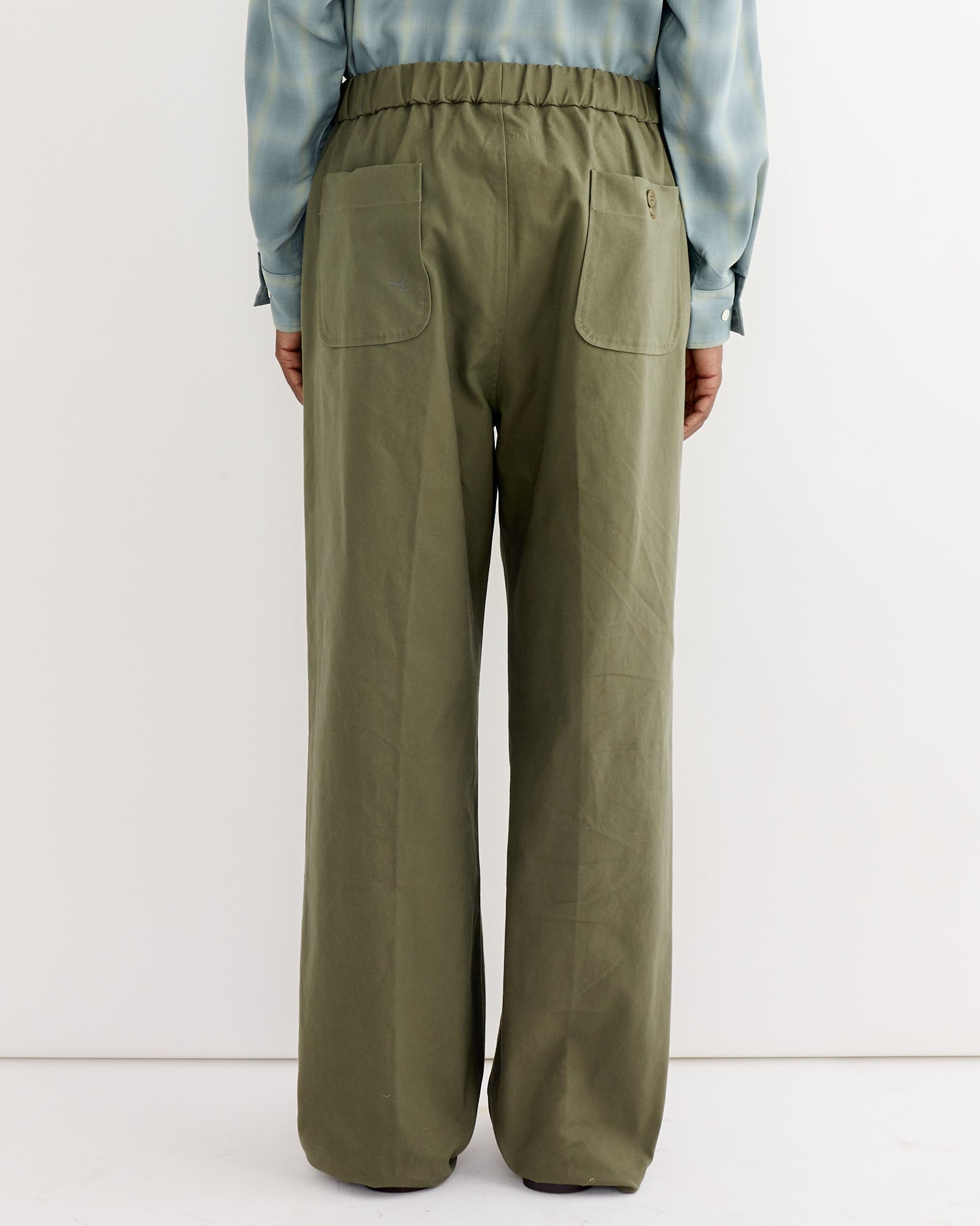A person stands with their back to the camera, wearing the SMOCK Kogai Pant in Duck Canvas Olive and a light blue long-sleeve shirt against a plain white background.