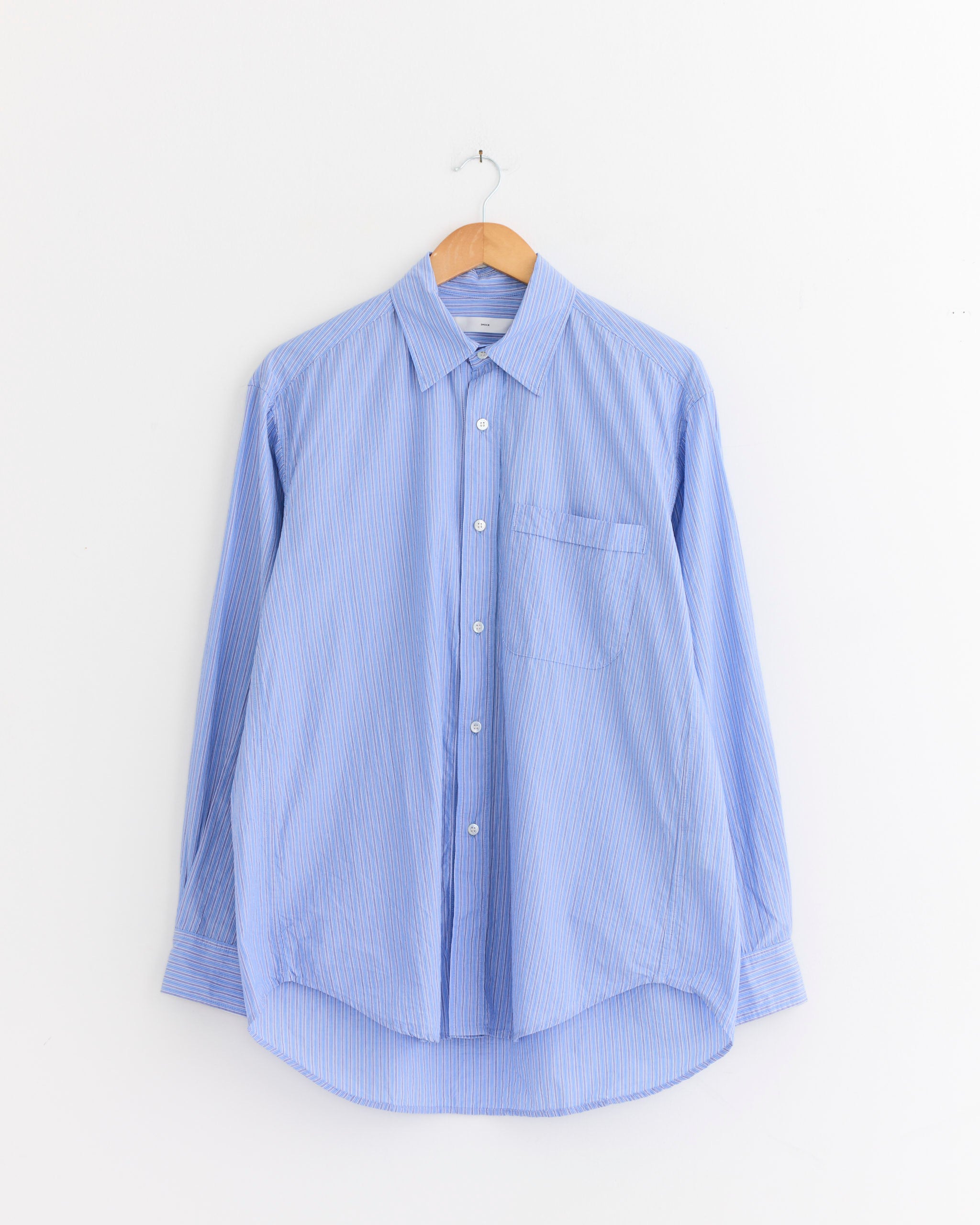 The SMOCK Gio Shirt in Crushed Cotton Blue, featuring white vertical stripes and wrinkle-resistant Japanese cotton, hangs on a wooden hanger against a white wall—a stylish travel-ready button-up shirt.