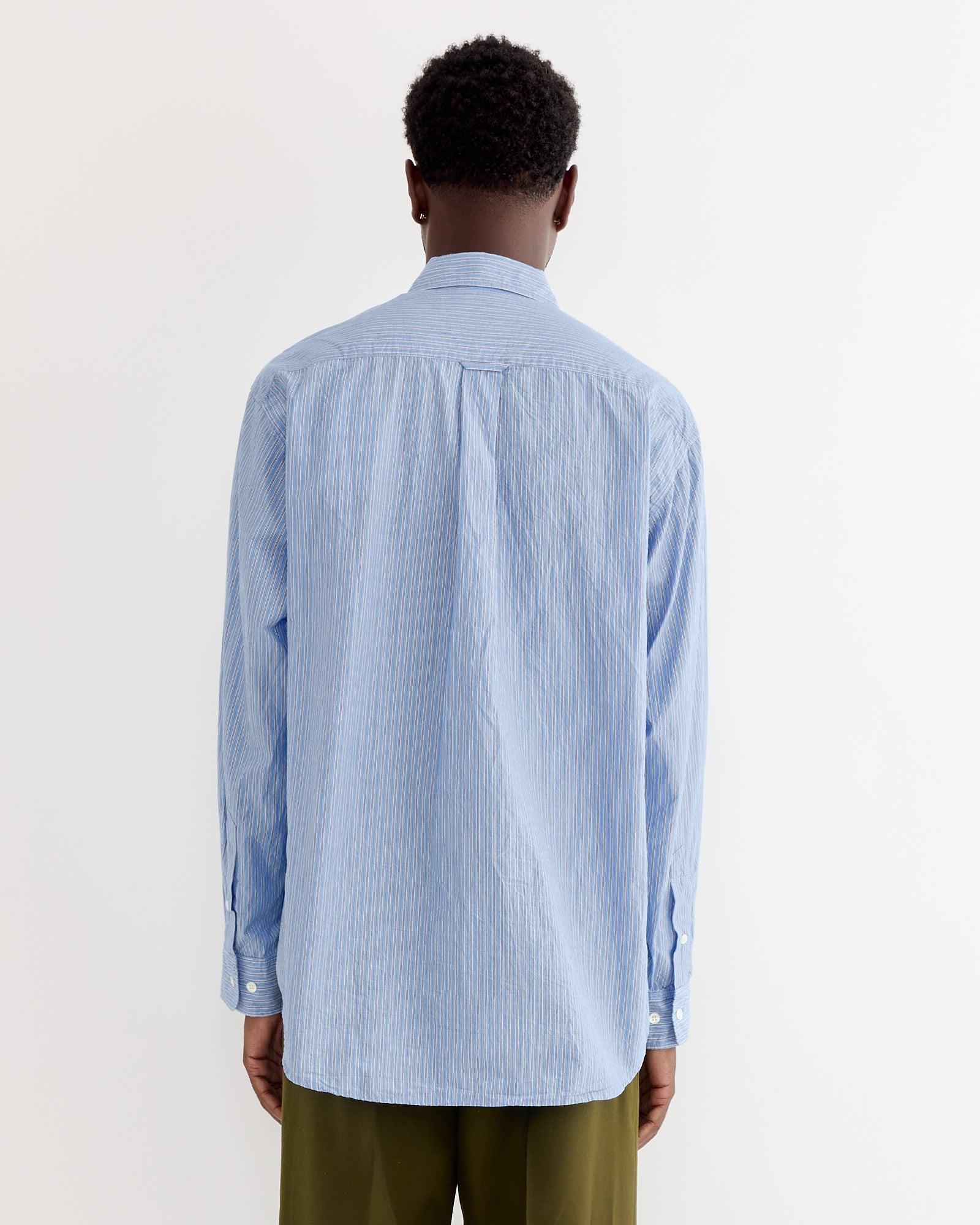 A person with short curly hair, wearing the SMOCK Gio Shirt in Crushed Cotton Blue Stripe Ni and olive green pants, stands with their back to the camera against a plain white background.
