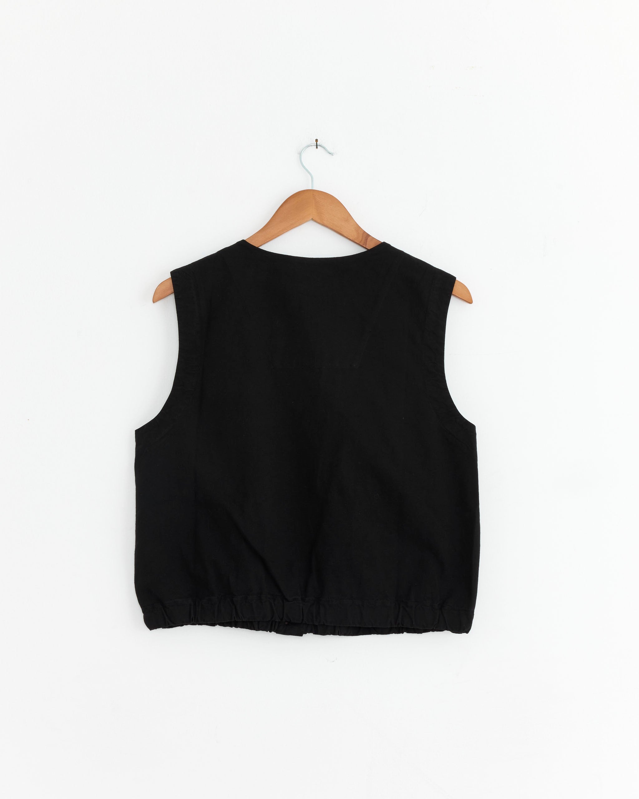 The Girls of Dust Veston Vest in Black, a sleeveless cropped style with an elastic hem, hangs on a wooden hanger against a plain white wall, displayed from the back.