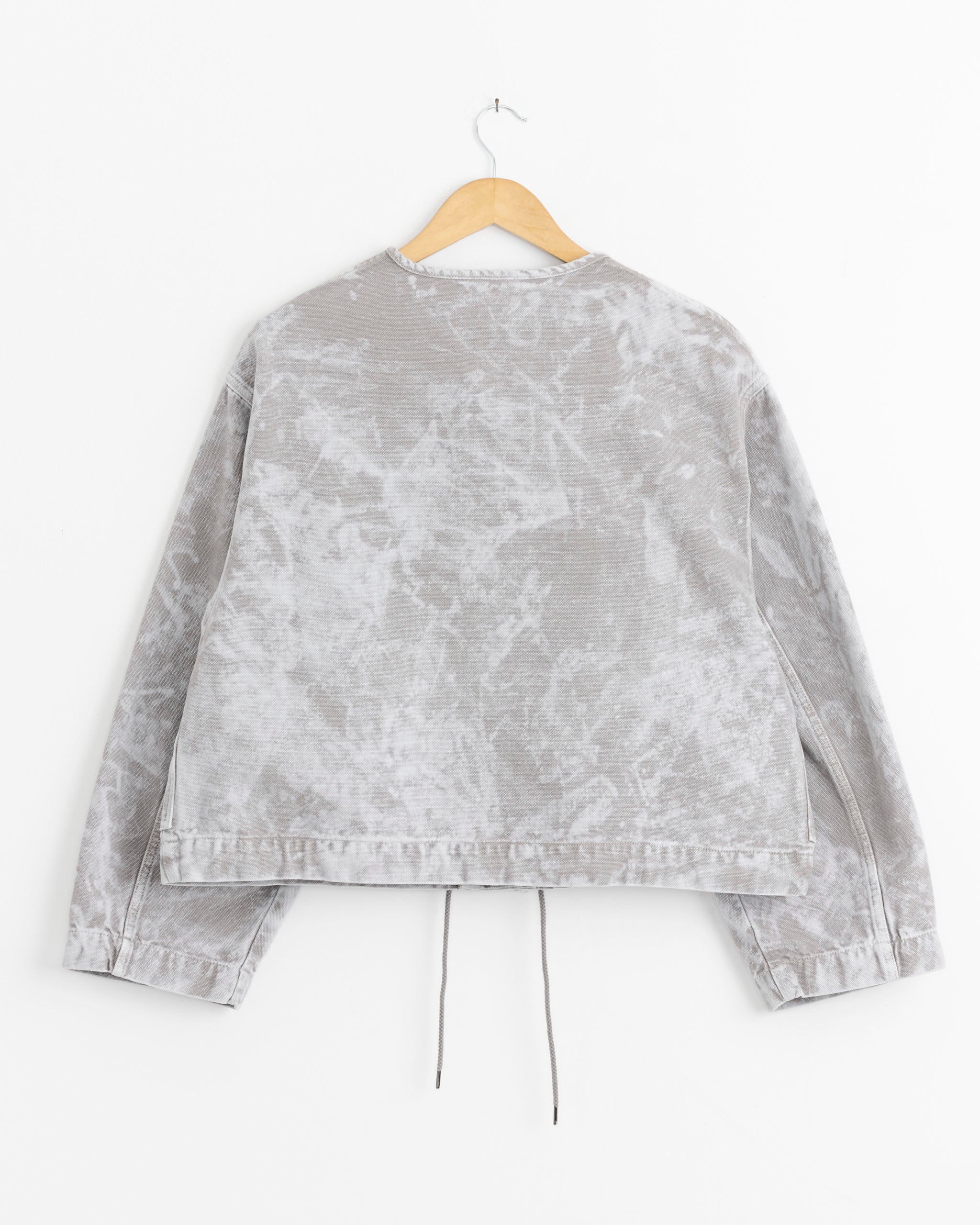 The Girls of Dust Nuclear Jacket in Marble is a light gray, long-sleeve, boxy fit jacket with a washed, marbled texture. It hangs on a wooden hanger against a white background and features a back hem drawstring for added detail.