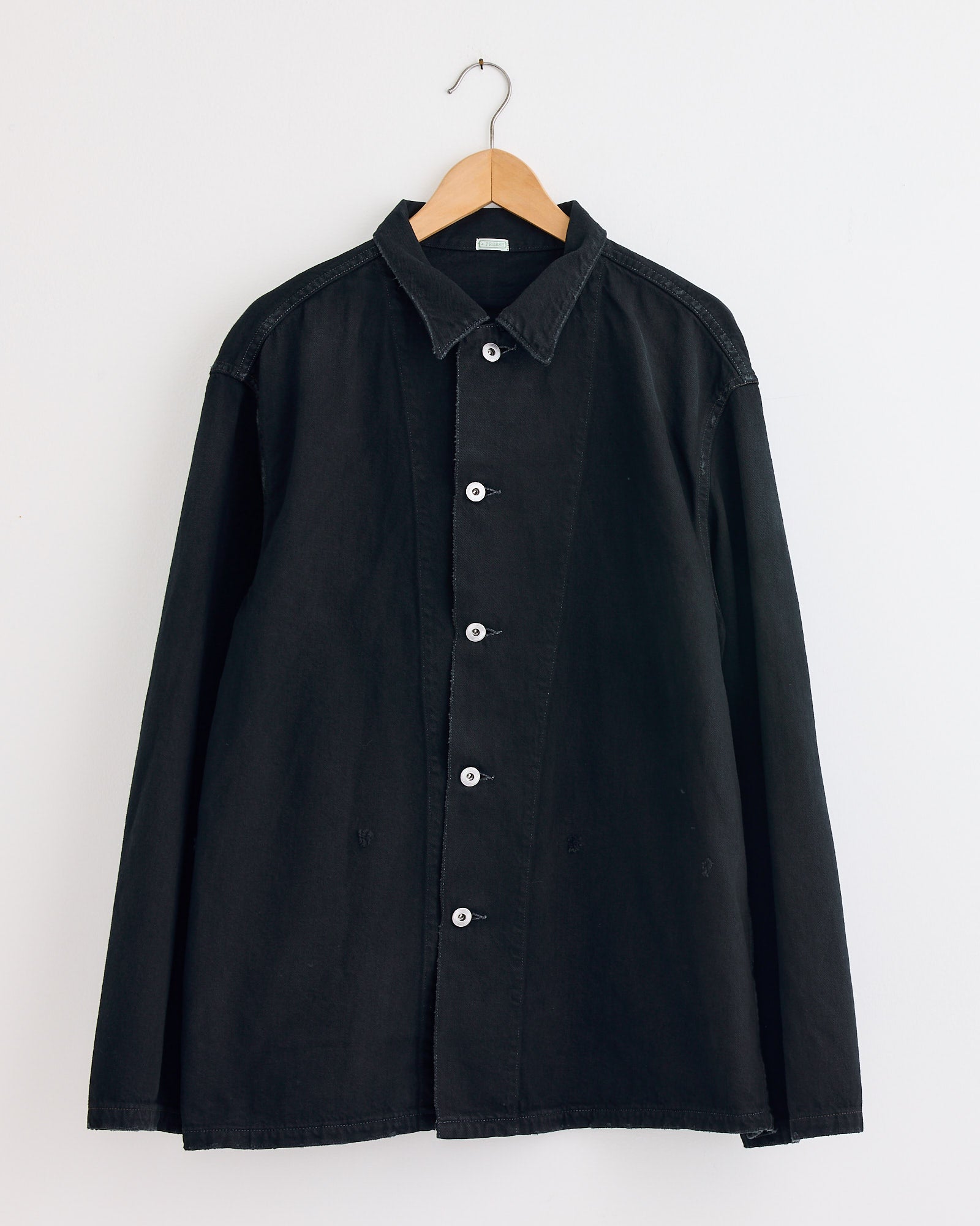 The A.PRESSE WW2 US Army Vintage Coverall in Custom is a black, collared, long-sleeve button-up shirt with white buttons. Crafted from vintage coverall fabric, it hangs on a wooden hanger against a plain white wall for a timeless look.