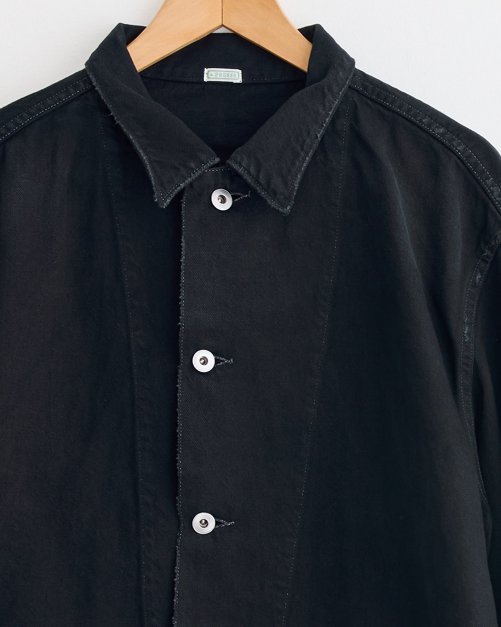 The A.PRESSE WW2 US Army Vintage Coverall in Custom is a black button-up shirt with a pointed collar, white buttons, and subtly frayed edges, displayed on a wooden hanger against a white background.