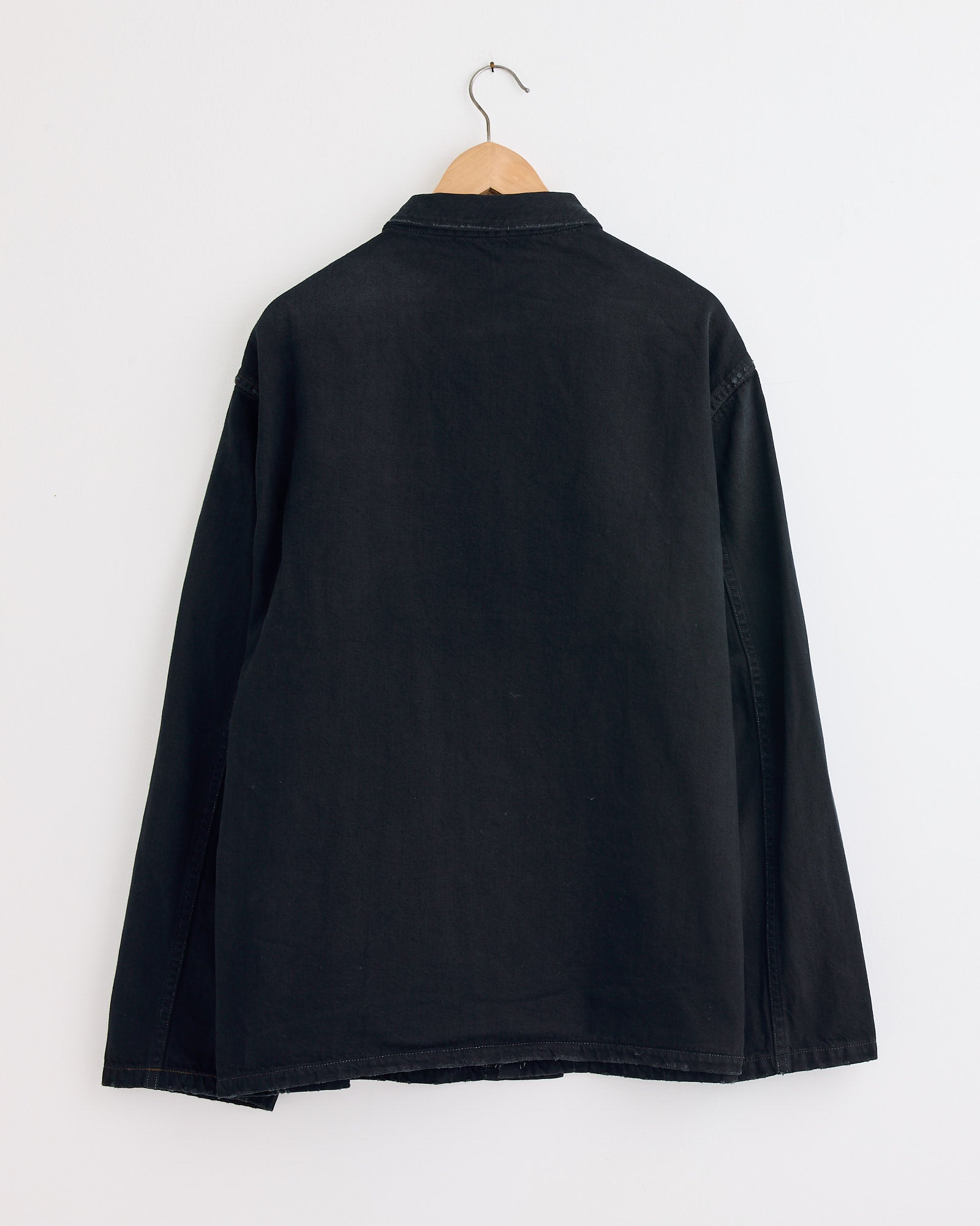 The A.PRESSE WW2 US Army Vintage Coverall in Custom, a black long-sleeve piece inspired by classic military designs, hangs on a wooden hanger against a white wall with the back view displayed.