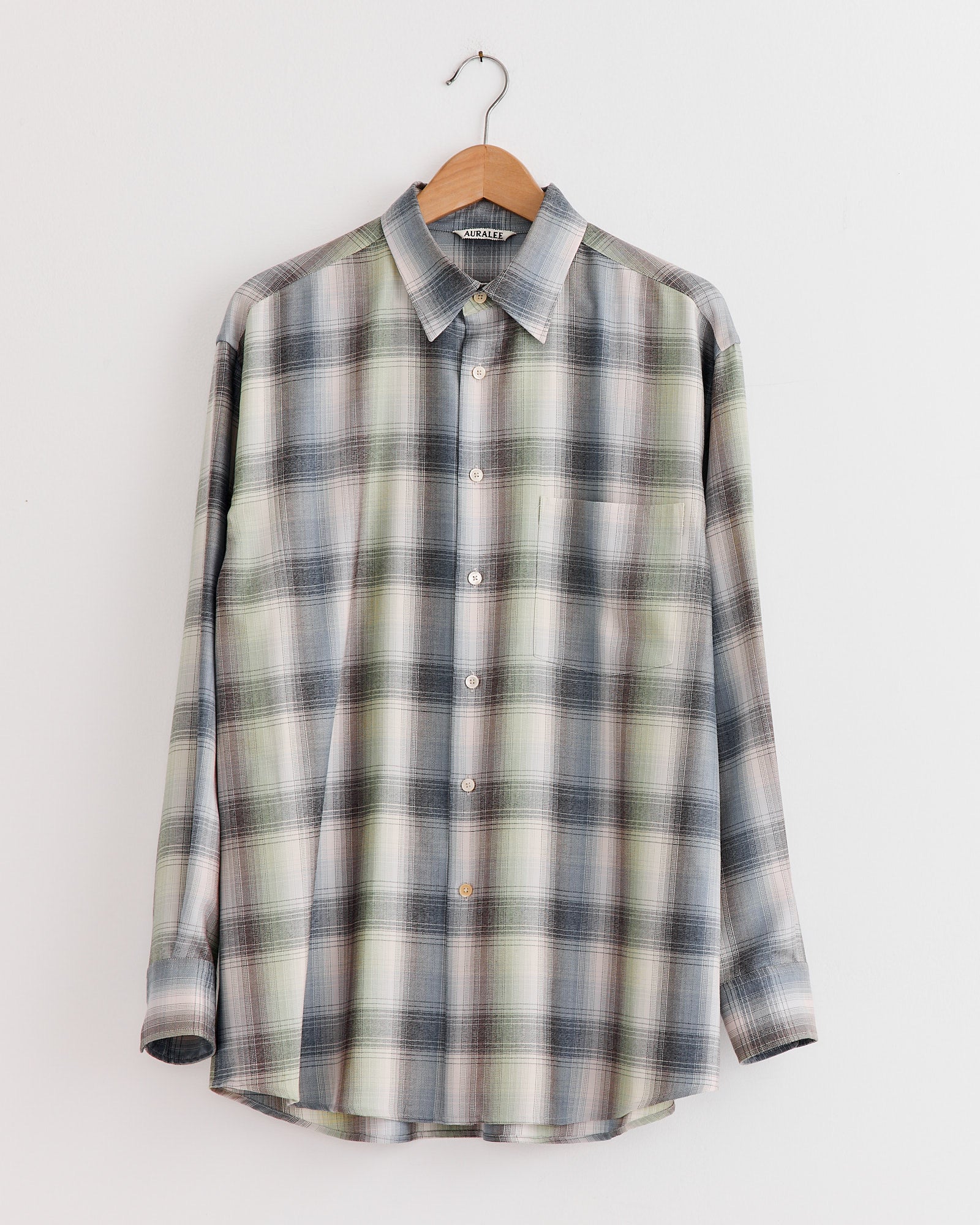 The Auralee Light Wool Check Shirt in Blue Green Check, a long-sleeve style, hangs on a wooden hanger against a plain white wall.