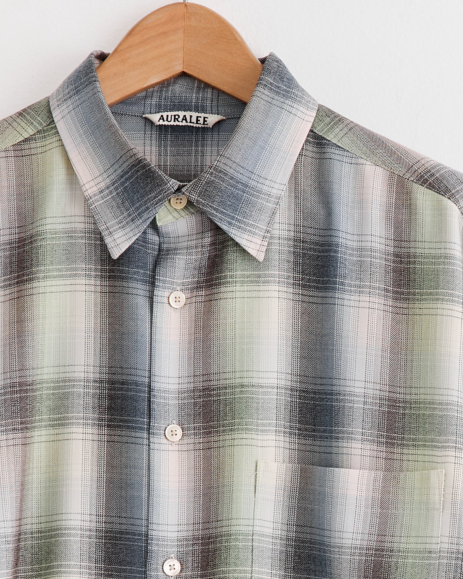 A close-up of the Auralee Light Wool Check Shirt in Blue Green Check, made from Super 120’s worsted wool with a pointed collar, beige buttons, chest pocket, and signature Auralee label, hangs on a wooden hanger.