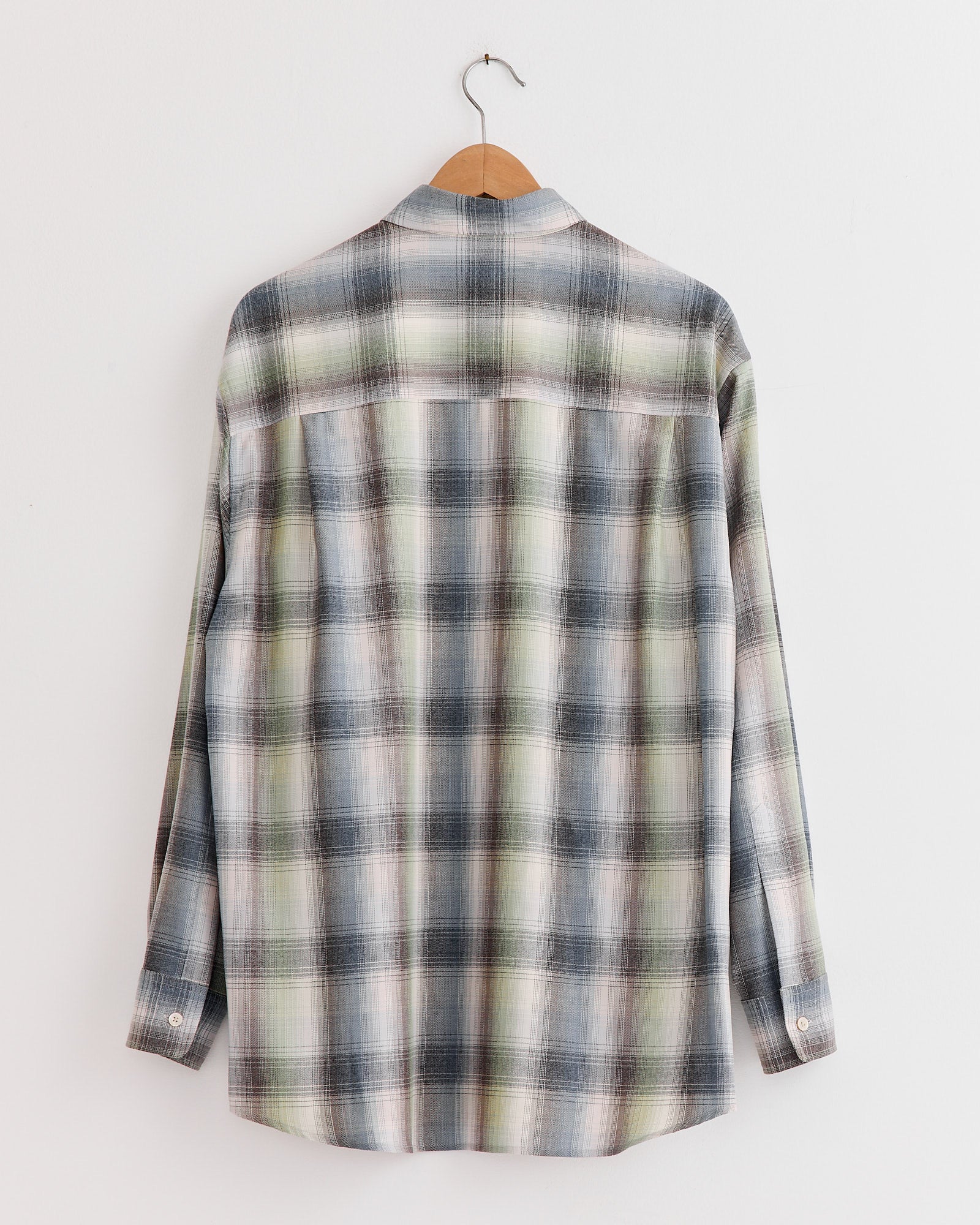 An Auralee Light Wool Check Shirt in Blue Green Check with long sleeves hangs on a wooden hanger, displayed back-facing against a plain white wall.